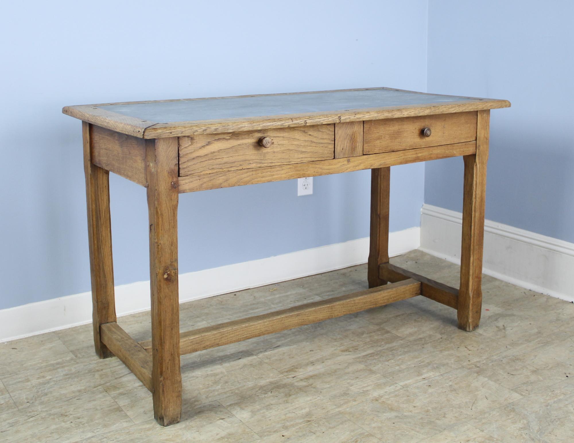 A solid oak writing table with a light gray zinc top. Trestle base is comfortable for resting one's feet. The carved F on the leg (shown in image #4), as well as the colorful interior of the drawers, are evidence of this tables past life in a French