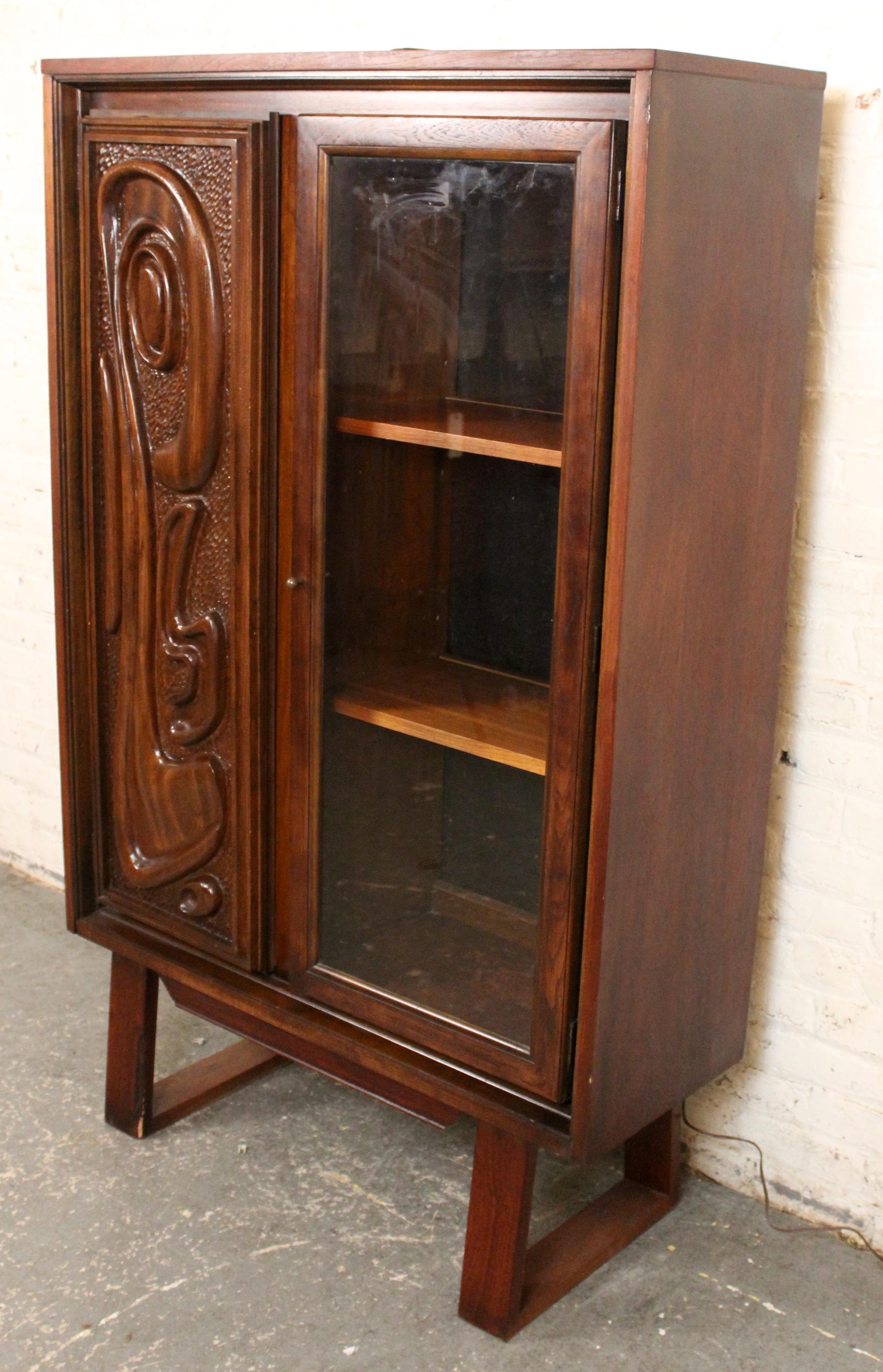 Dieser kühne und kunstvolle Mahagonischrank im Vintage-Stil von Pulaski zeichnet sich durch eine auffällige, skulpturale Fronttür mit abstrakten, ozeanisch anmutenden Schnitzereien aus. Die gegenüberliegende Tür ist mit einer Glasscheibe versehen,