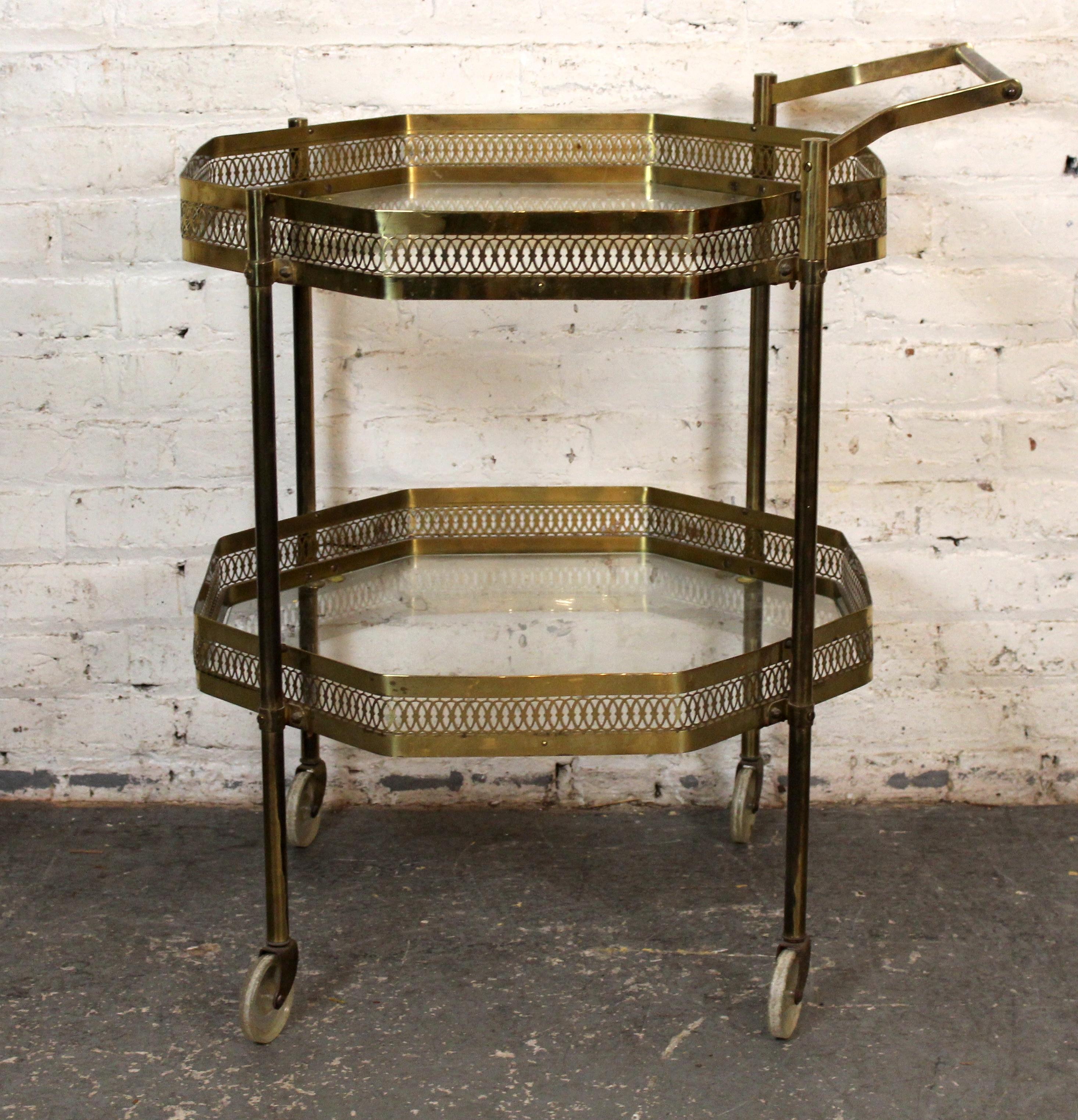 Chic and sculptural, this octagonal brass bar cart showcases a unique geometric frame with two tiers of clear glass shelving. The warm brass finish exudes vintage sophistication. Set on fixed wheels for smooth mobility, it's a stylish and functional