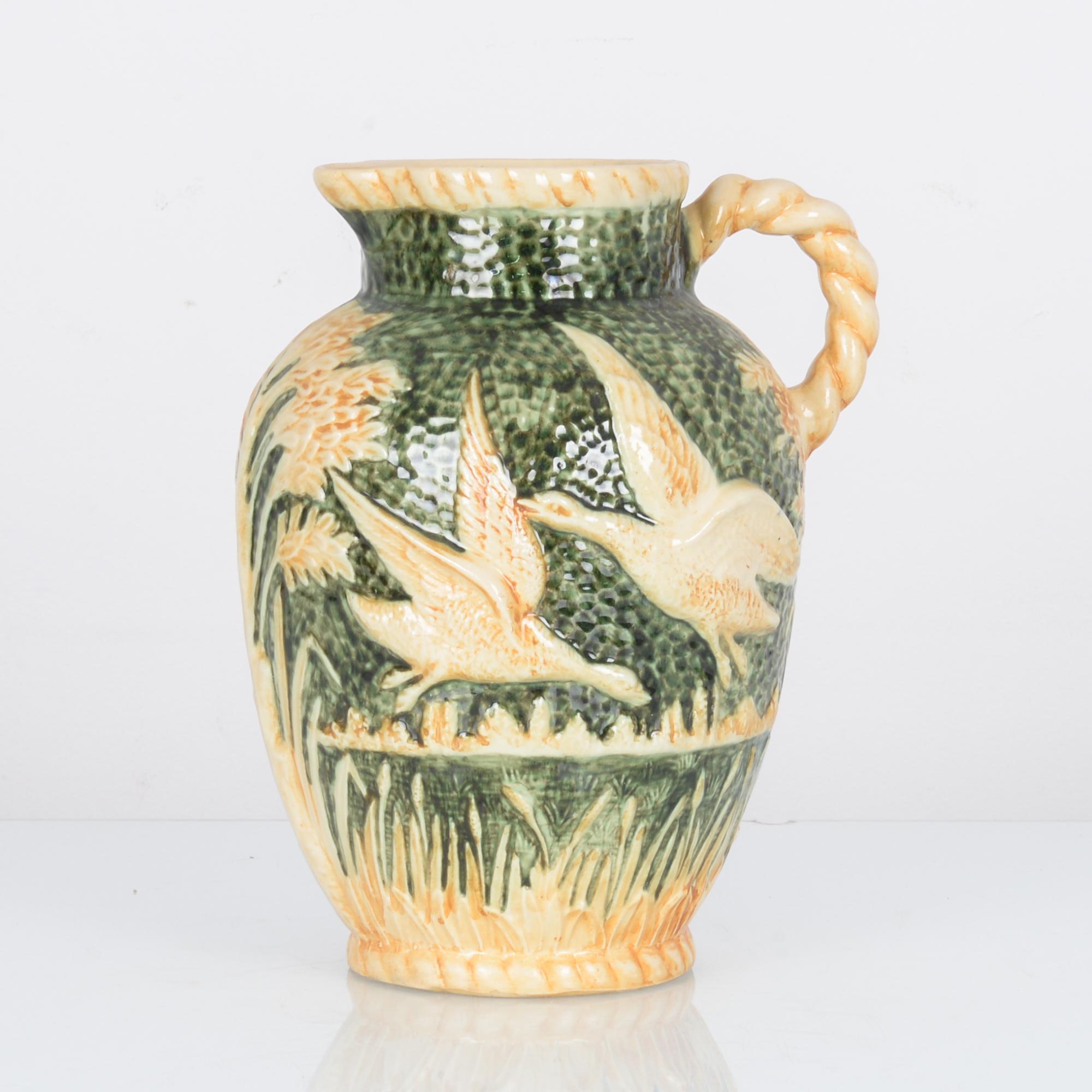 A green/tan ceramic vase with handle, produced in Belgium circa 1960. A 30 cm ceramic with a scene of flying birds carved in relief. Dimpling under the rich green glaze brings the image to life. The twisting handle calls to mind reeds freshly picked