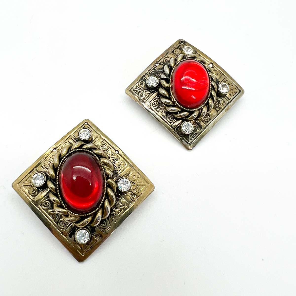 A wonderful pair of Vintage Silver & Ruby Plaque Earrings. Chased panelling set with a rope twist design is topped with a large ruby red cabochon and a quad of mounted white chatons. A gloriously glamorous finish to your look.

Vintage Condition: