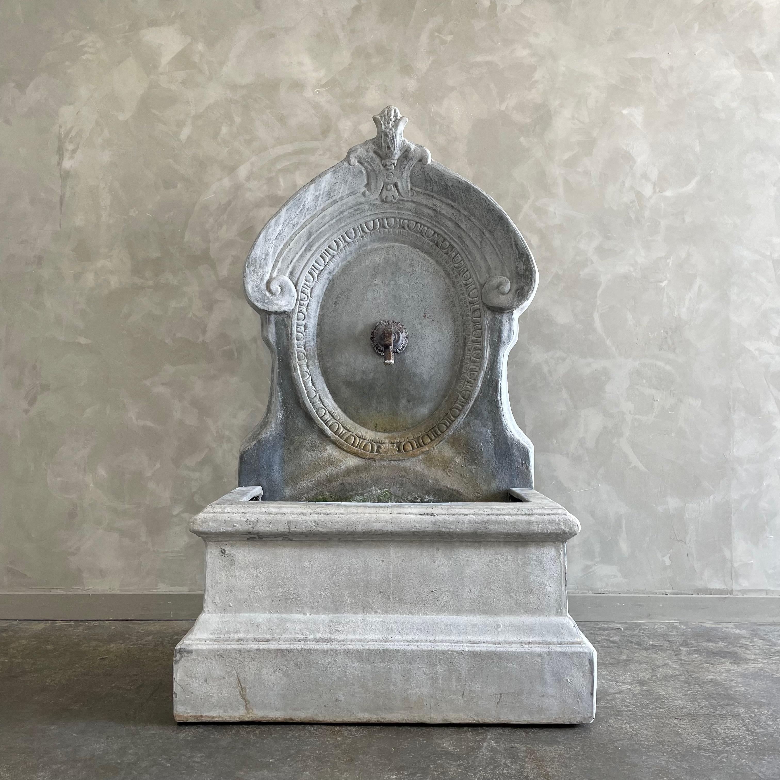 Fontana da giardino rustica vintage ispirata alla scultura francese e all'artigianato minimalista. La fontana in pietra e fibra di vetro e il beccuccio in ferro spruzzano acqua con un sereno rumore bianco.