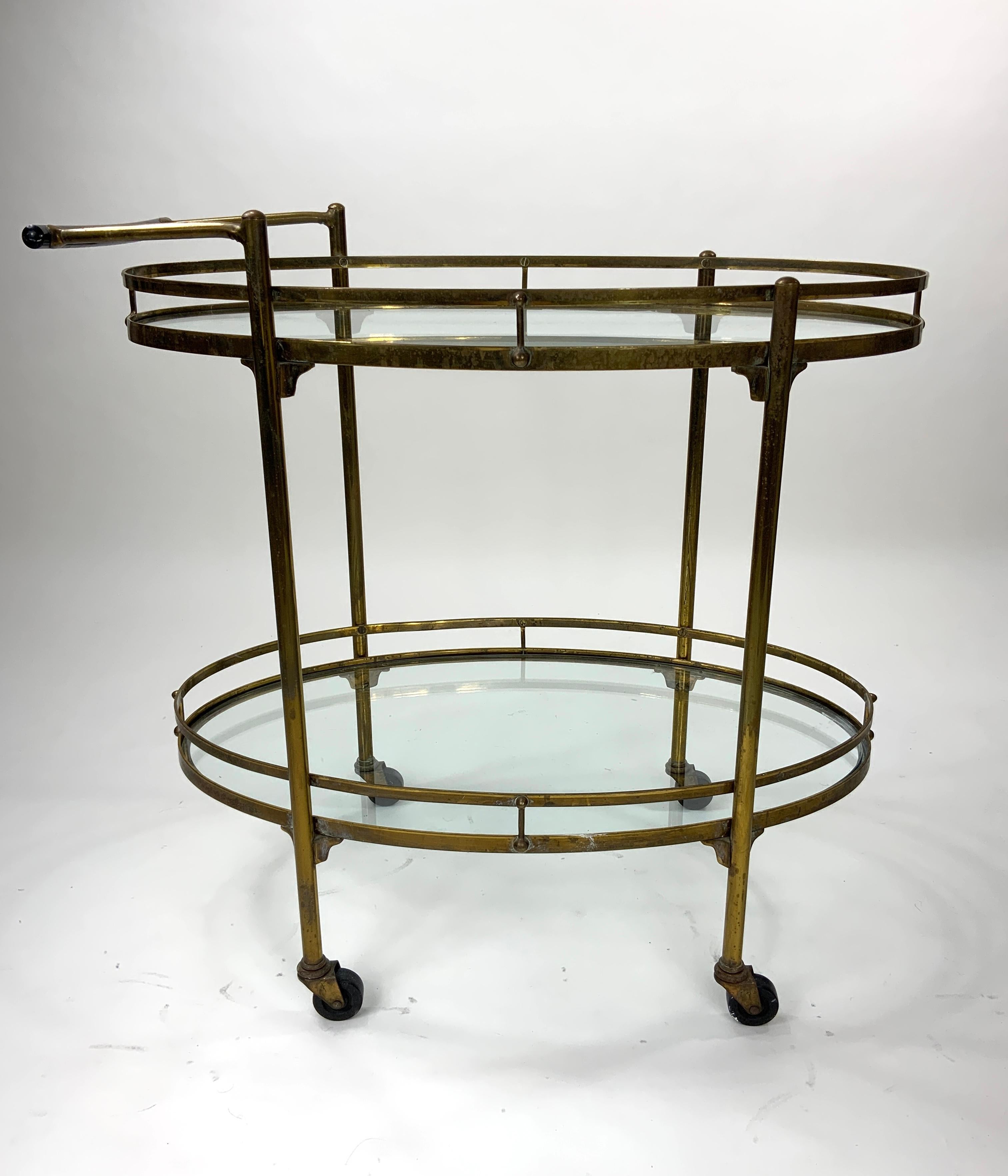 An oval brass bar cart/tea trolley from the 1940’s and possibly late 1930’s attributed to barware and furniture manufacturer Maxwell Phillips of New York. It has 2 tiers with each having a double-ribboned guardrail and a glass shelf. The cart sits