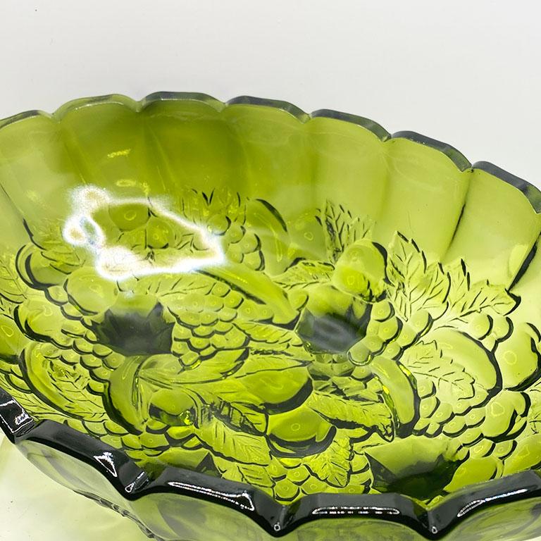 A transparent green glass fruit bowl or serving platter. With scalloped edges, and a textured fruit motif, this vintage bowl sits upon four raised glass legs. The edges are scalloped and in an oval shape. Set this piece on a kitchen counter to