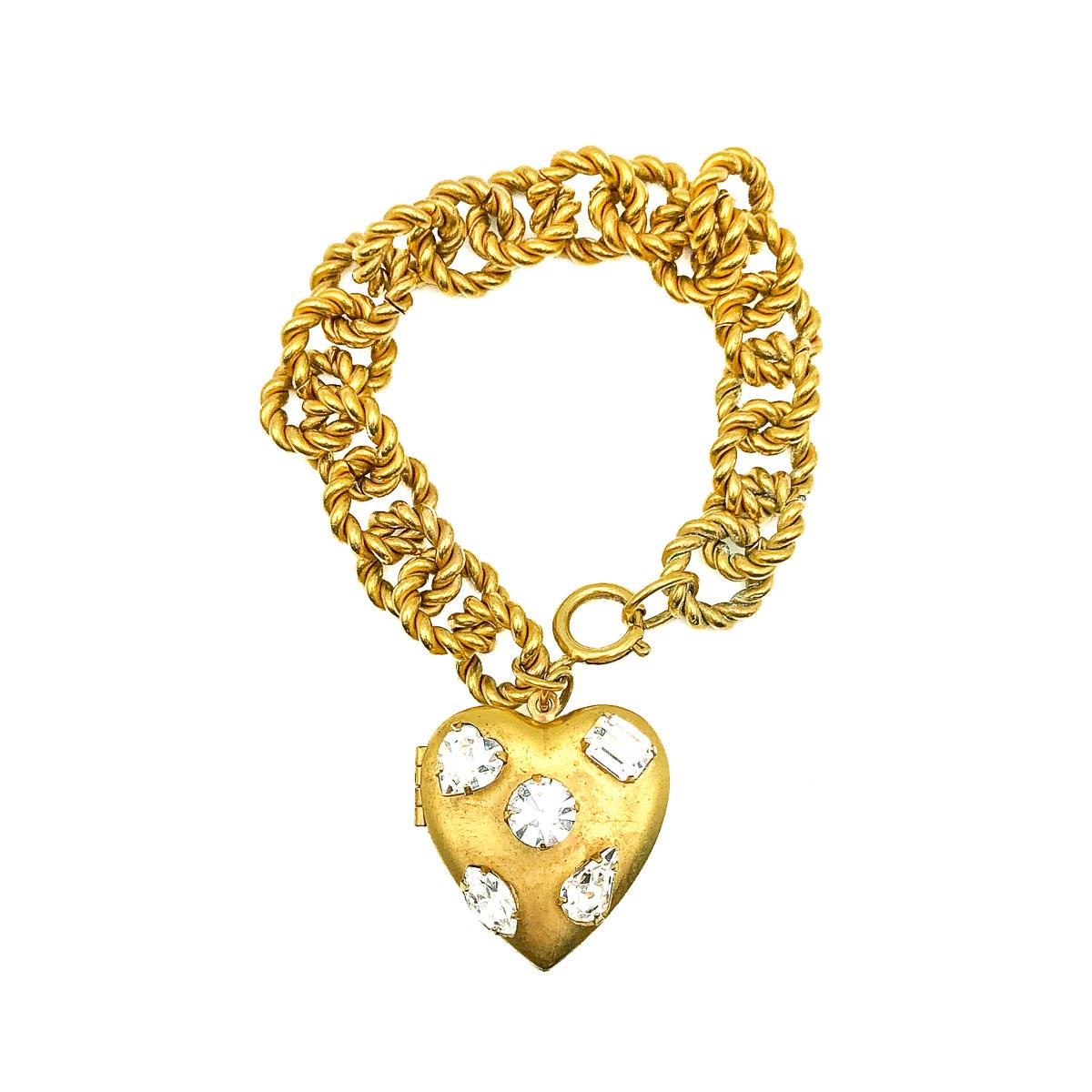 Un bracelet médaillon en forme de cœur avec une attention particulière aux détails. Réalisé en métal plaqué or et pierres de cristal de taille fantaisie. Un médaillon à grande ouverture en forme de cœur est orné de cinq pierres, chacune d'une taille