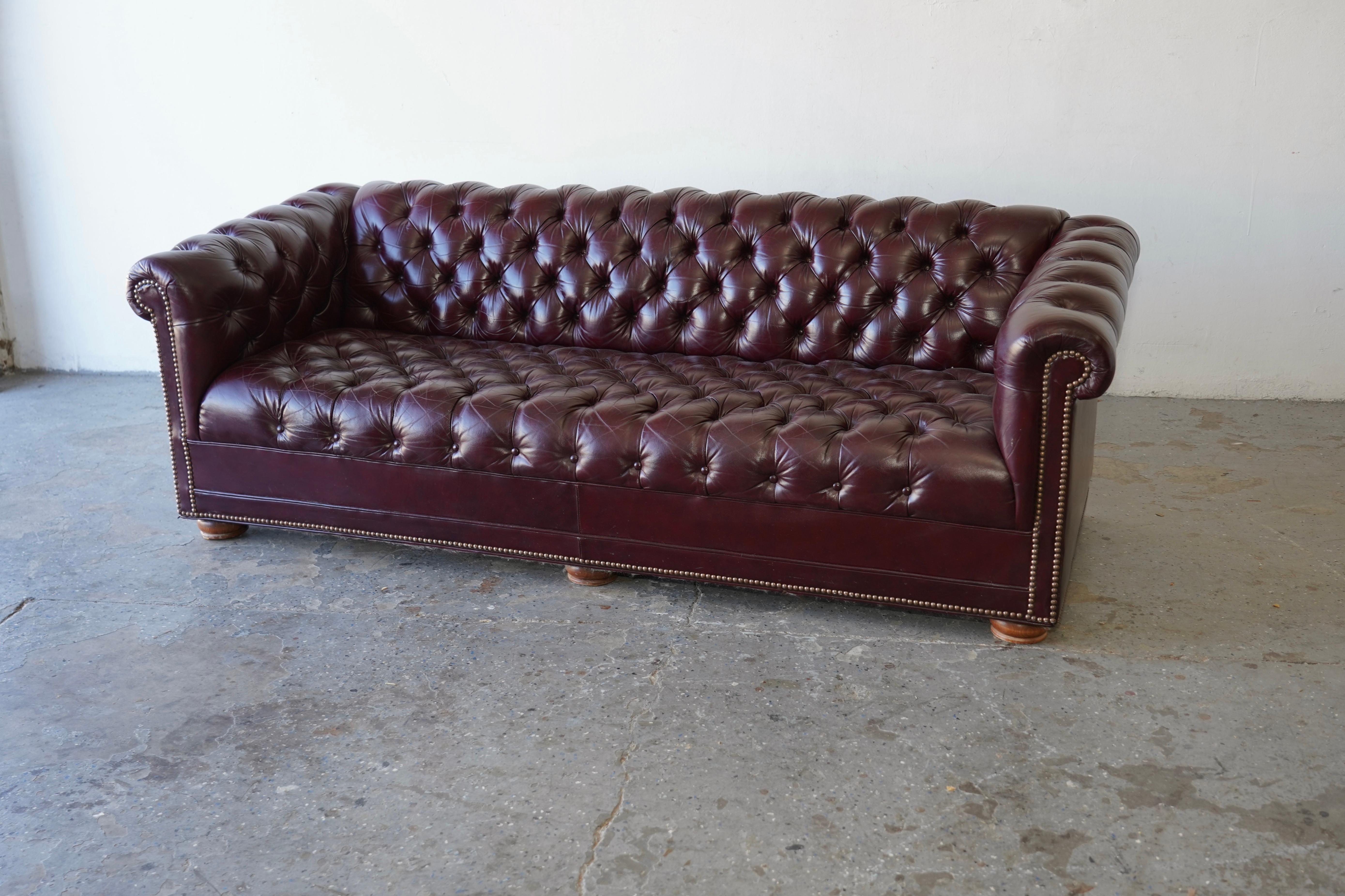 Stunning vintage tufted oxblood  leather  Chesterfield sofa, exuding timeless elegance. The rich, smoky burgundy hue adds depth and sophistication, truly coming to life when illuminated. Featuring classic bun feet and brass tack heads, this high-end