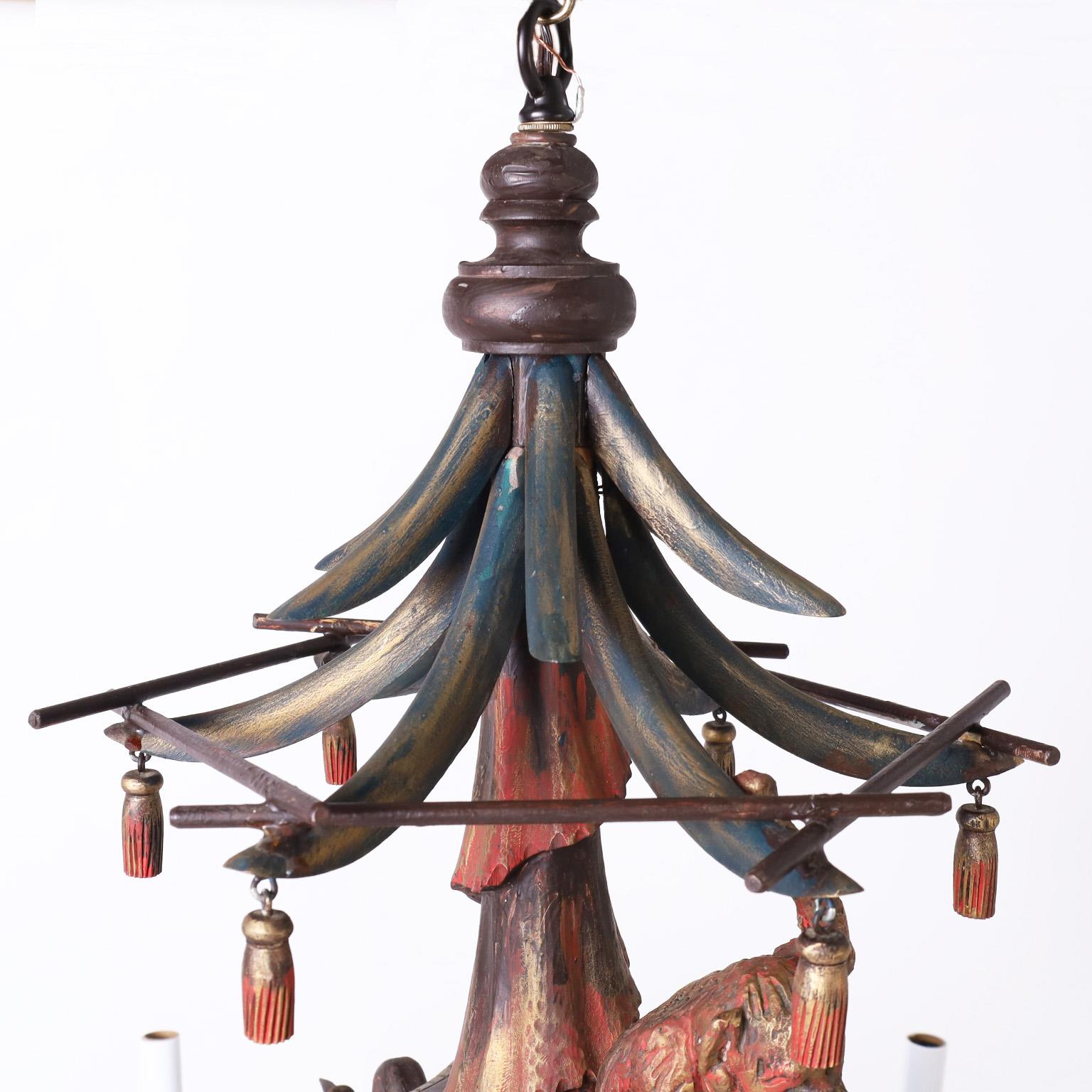 Whimsical vintage chandelier with asian motifs crafted in wood paint decorated in a polychrome style featuring a pagoda form top, carved drapery, wood tassels, a carved monkey, faux bamboo arms and supports with an artichoke finial. One of two
