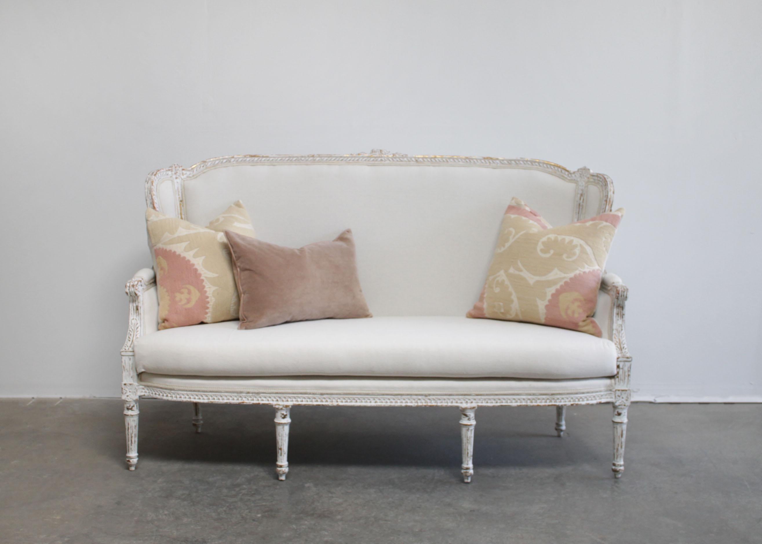 Vintage painted and upholstered Louis XVI style sofa settee
Painted in a french white with subtle distressed edges, where gilt and wood show through.
Classic fluted legs, with a medallion carving, and large flower carving at 
the top back of the