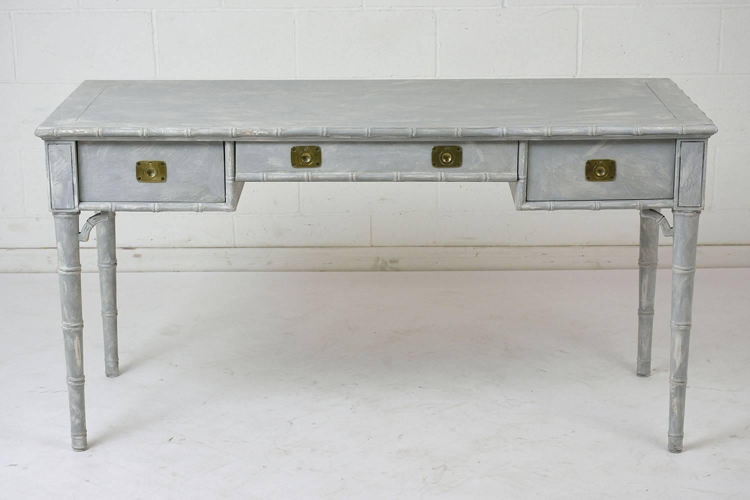 This 1970s modern-style desk is made of wood painted in a pale gray and oyster color combination with a distressed finish. The desk features moulding details carved to resemble bamboo. There are three drawers in the desk with flush brass drawer