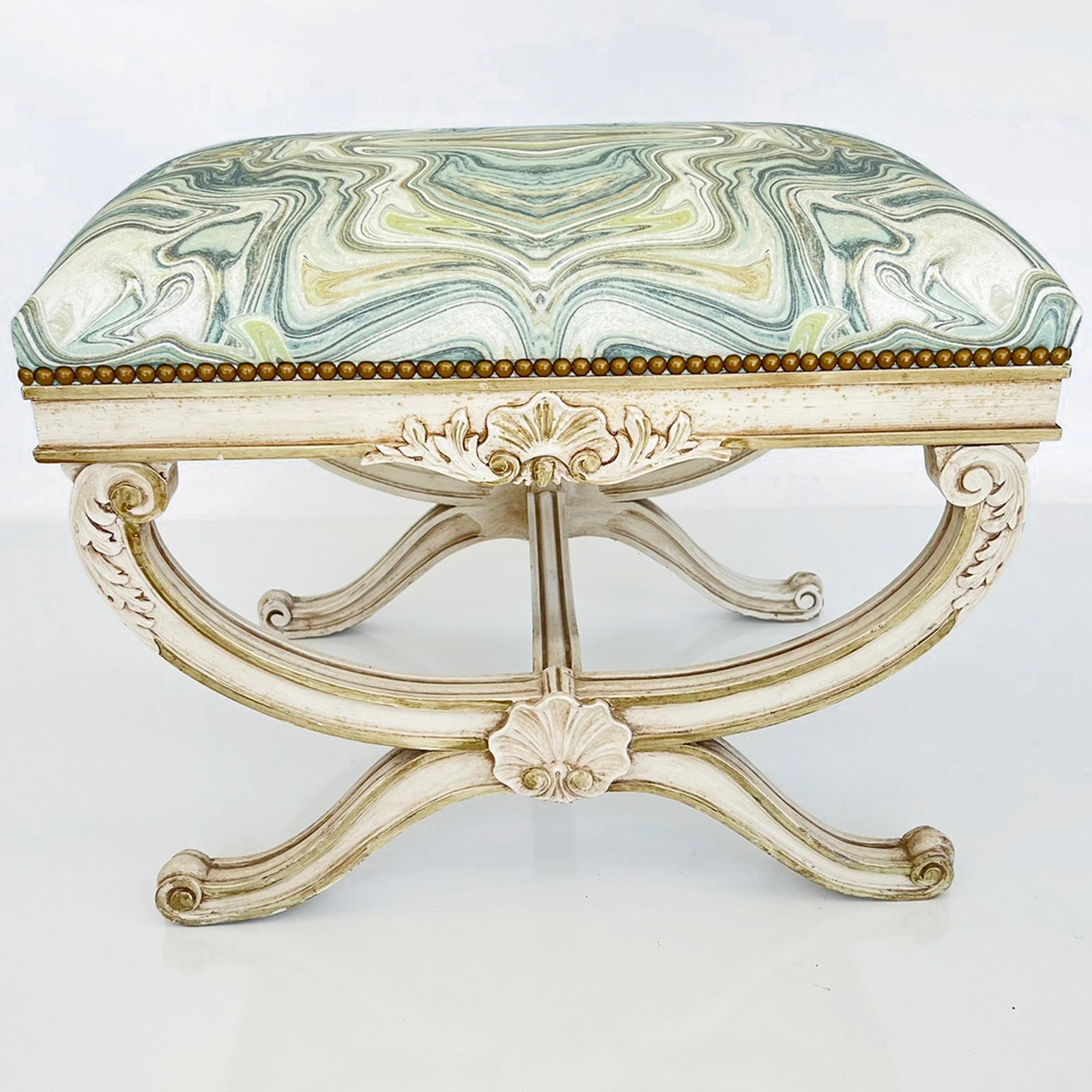 Curule-form bench, having a padded, crown seat of onyx-printed linen with nailheads, on an ivory painted frame showing natural wear, its fielded apron centered with an acanthus-flanked scallop shell, raised on channeled, acanthine-carved, X-frame