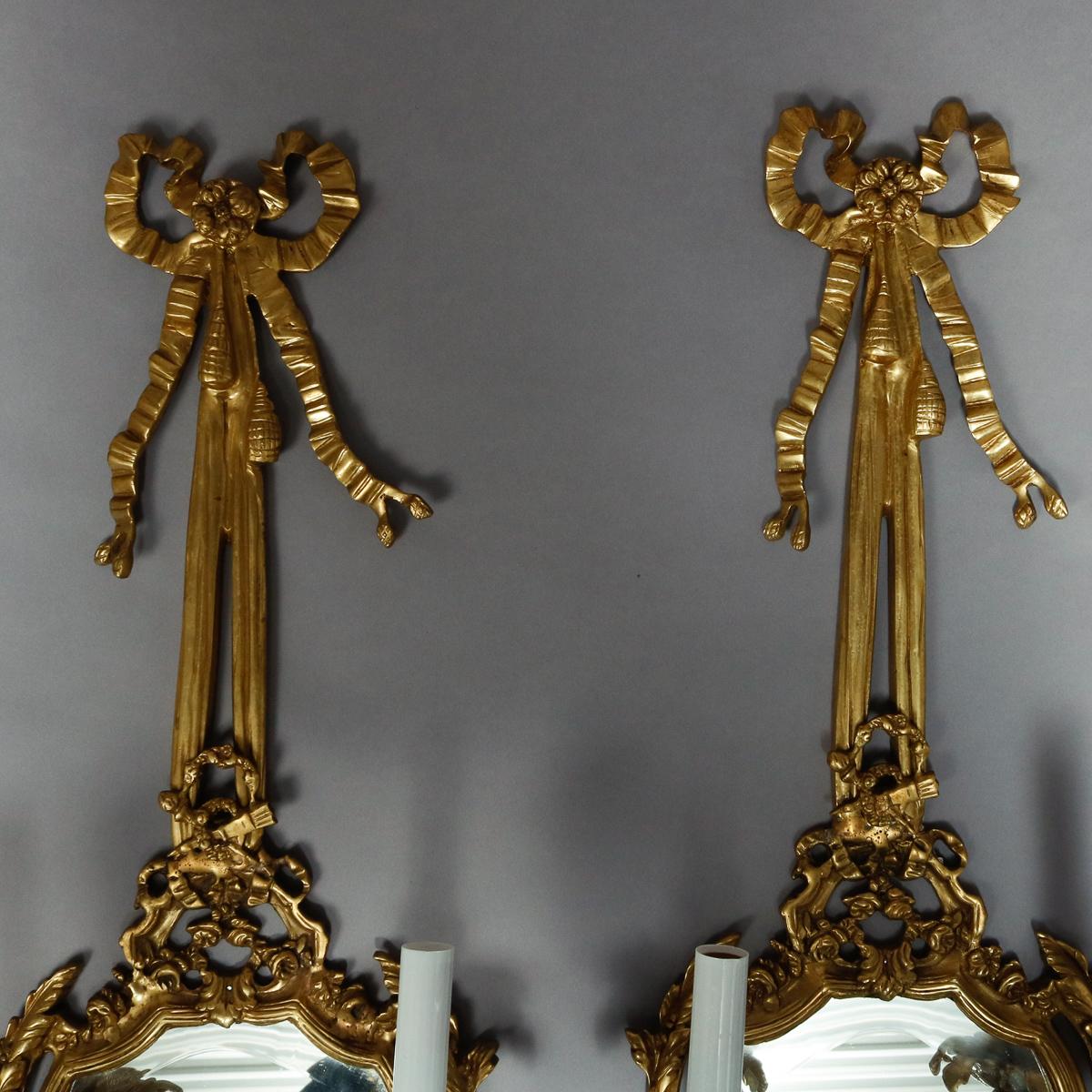 A vintage pair of electric wall sconces offer cast brass frames with tied bow form crest over shield shaped and foliate framed beveled mirror having two candle lights, 20th century

Measures: 31