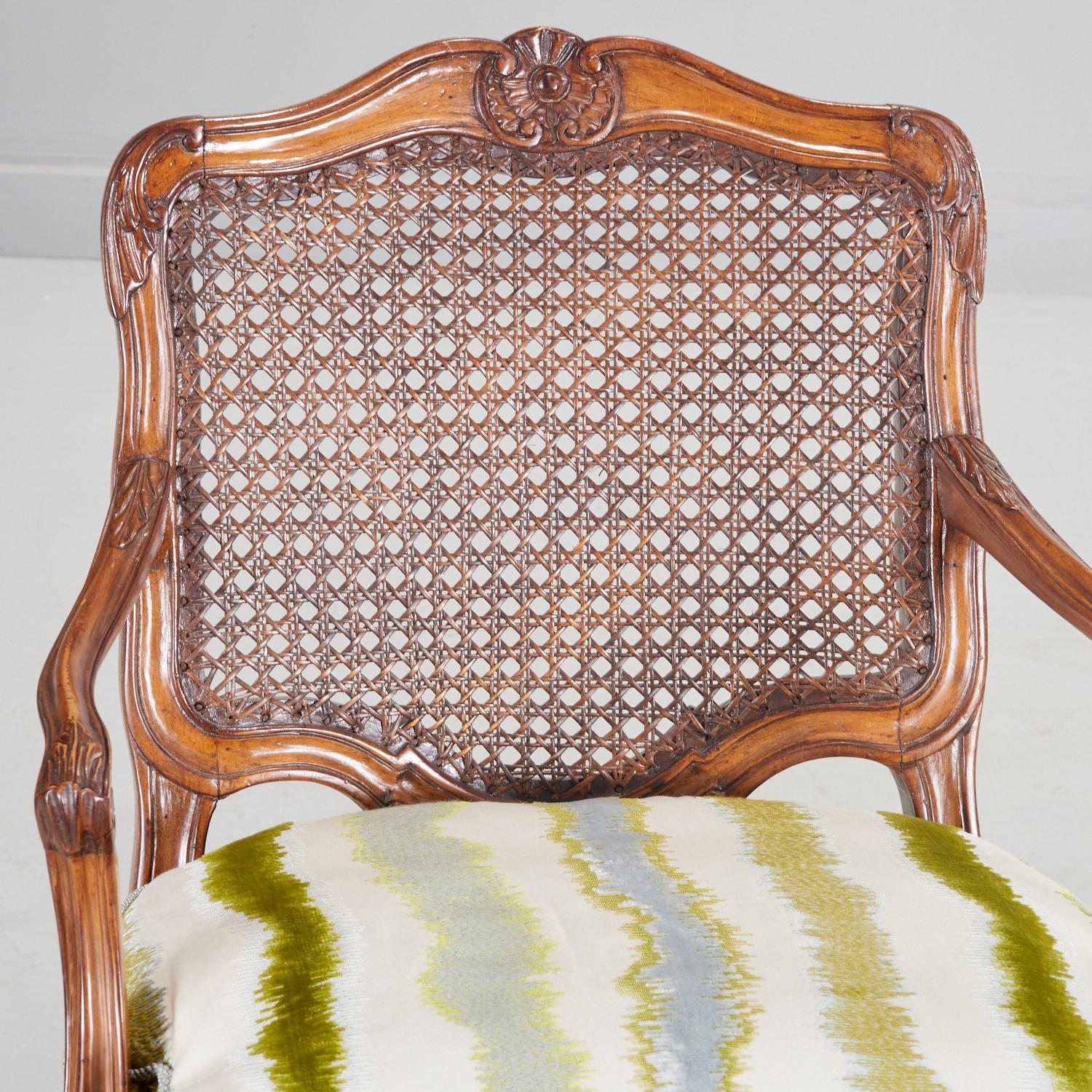 A pair of 20th century, Louis XV style carved fauteuils with caned back and upholstered seat. The beechwood channel carved frame is decorated with rosettes and acanthus fronds. Cabriole legs terminate in whorl feet. A loose down-filled seat cushion