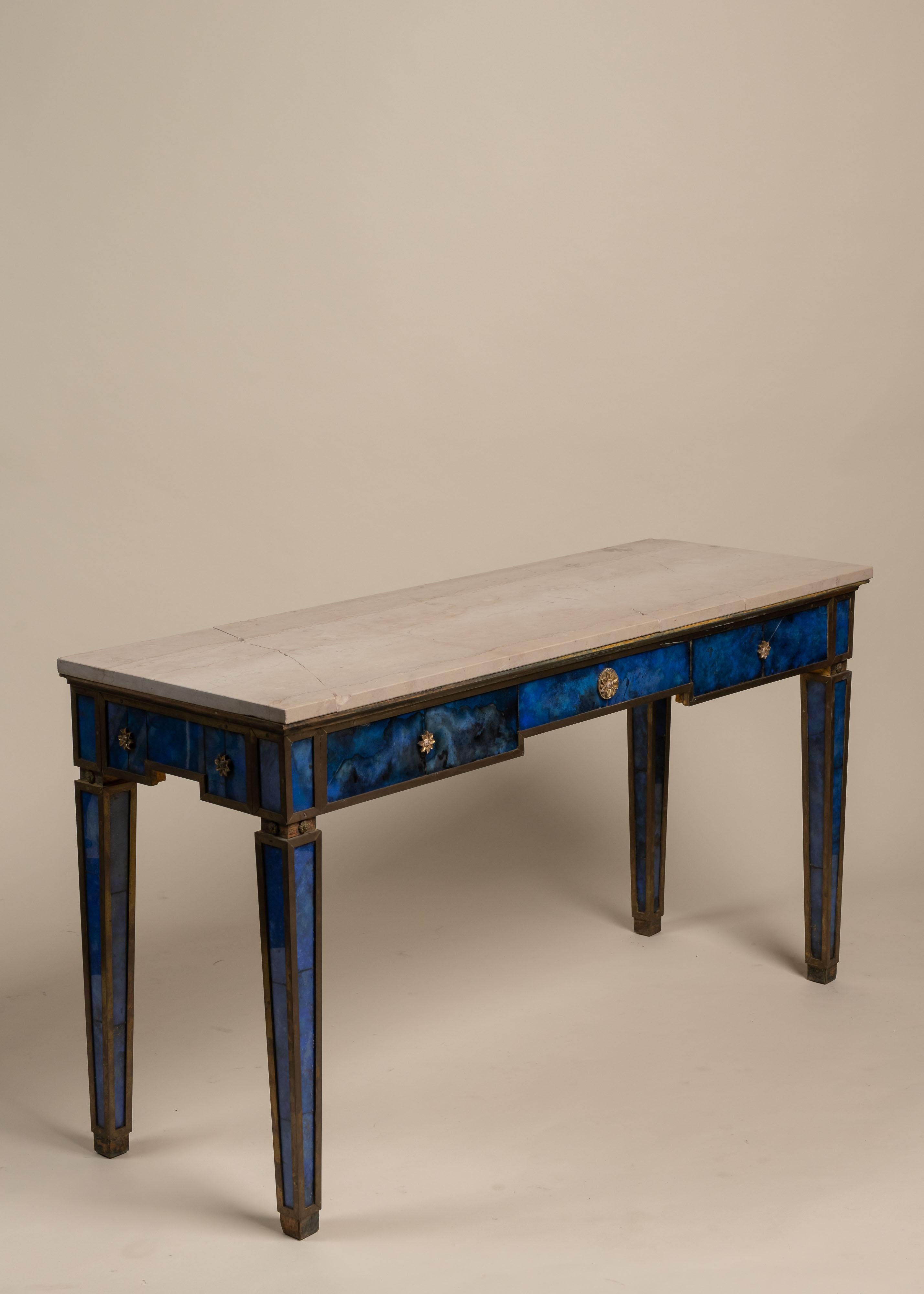 These refined console tables echo the neoclassical elegance of Maison Jansen. The clean lines and tapered legs are accented with panels of deep blue églomisé glass and gilt trim. Three drawers feature star-shaped gilt bronze rosettes, adding a