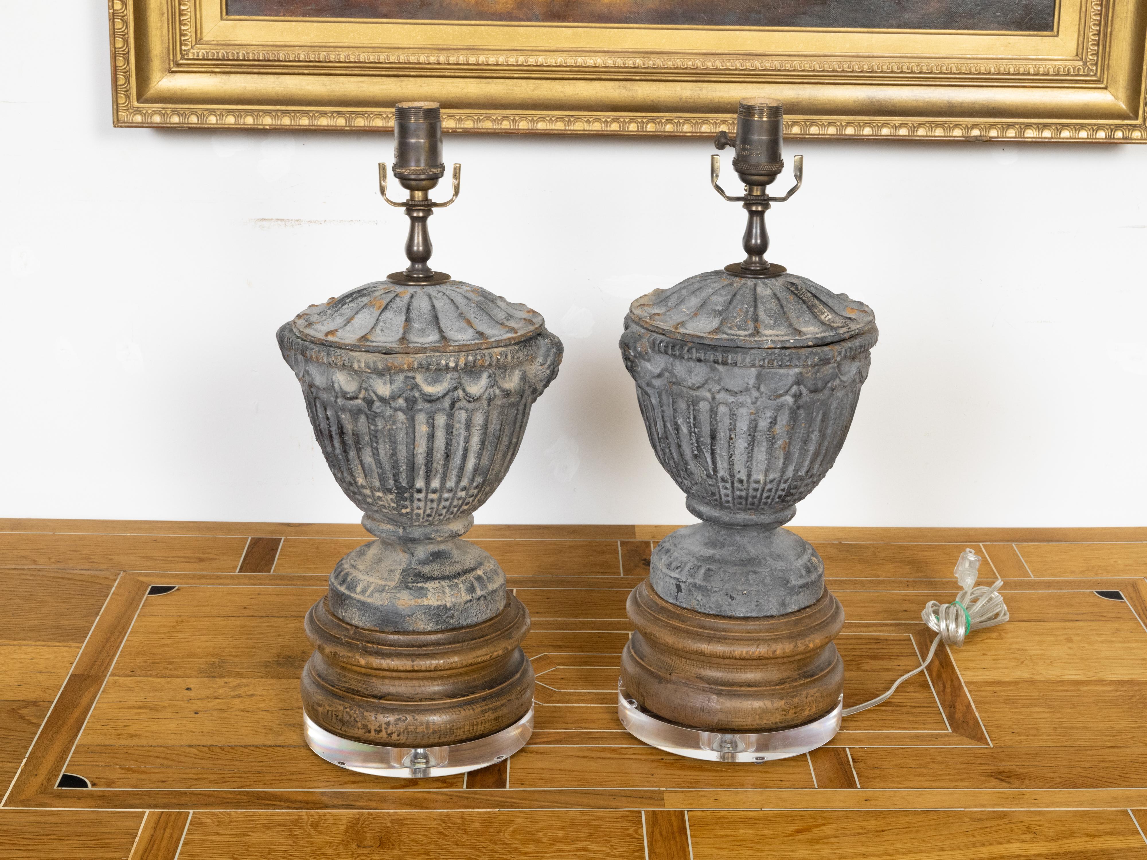 A pair of French iron table lamps from the mid 20th century, on wooden and lucite bases. Created in France during the Midcentury period, each of this pair of table lamps features an iron lidded vase adorned with fluted accents and petite decorative