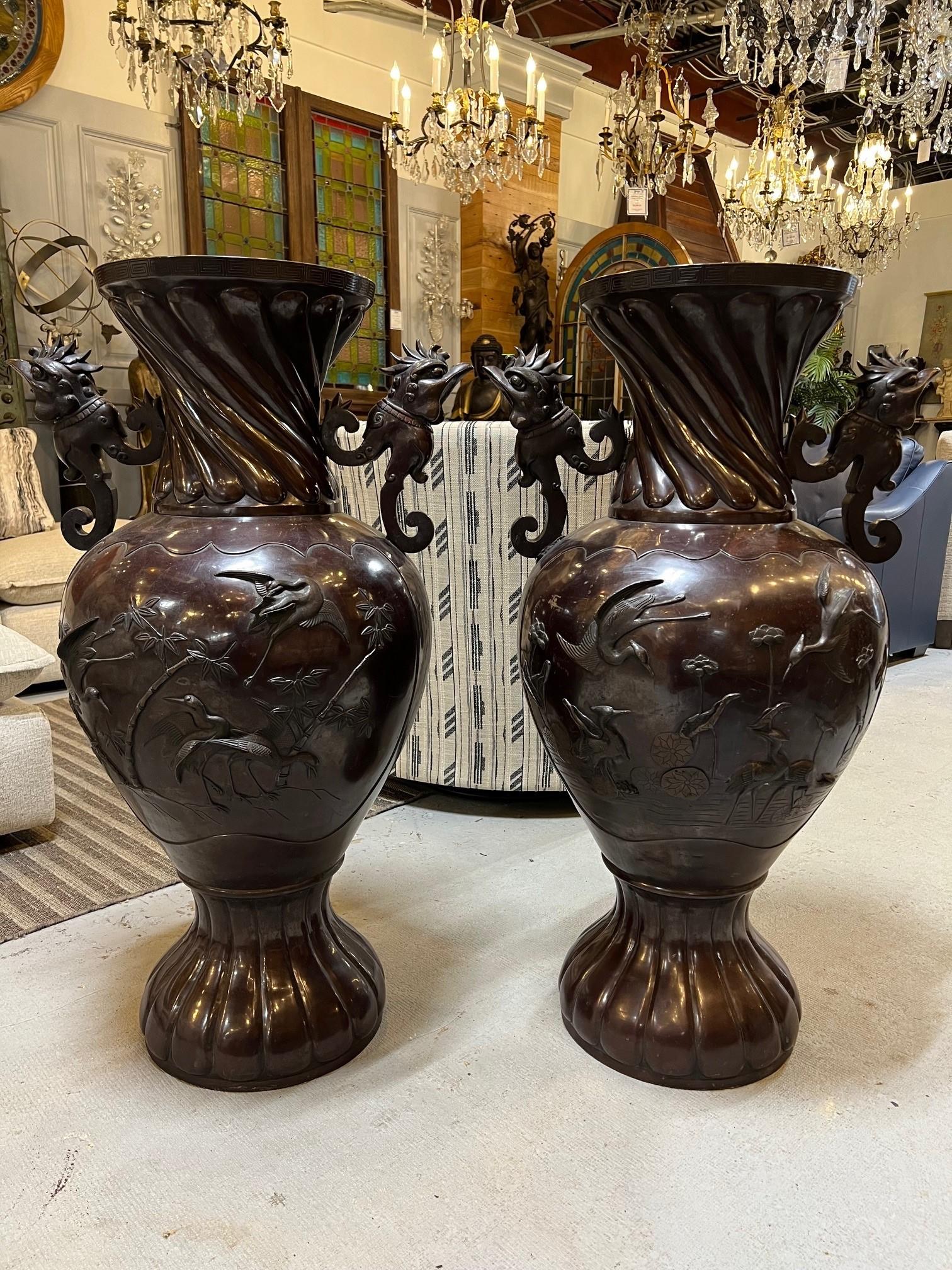 An attractive pair of Mid 20th Century Japanese large bronze urns. Bird head handles and decorative panels on both sides of raised birds, trees, and flowers makes this a beautiful pair of urns. The urns are in very good condition and could be used