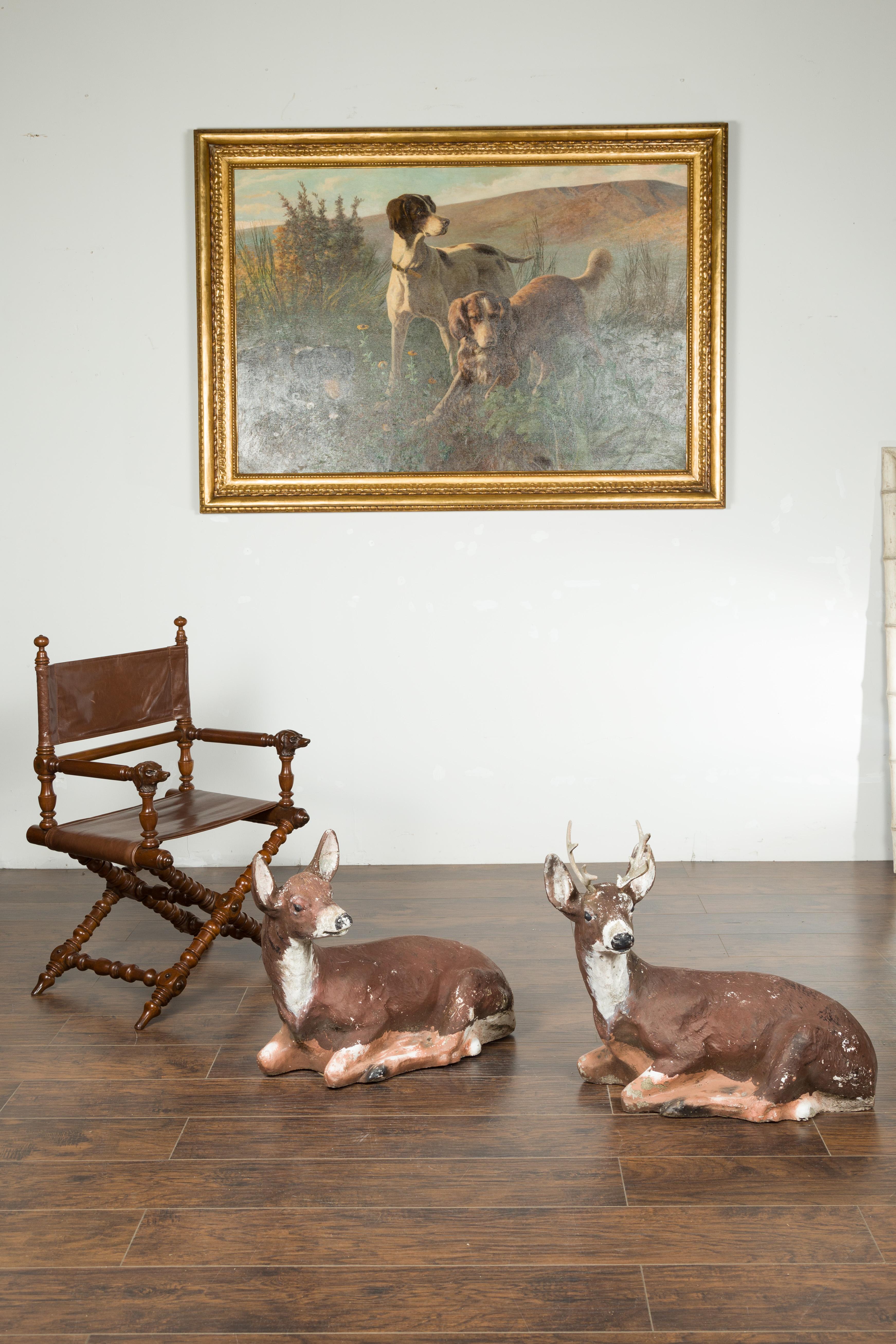 A pair of American painted concrete deer sculptures from the mid 20th century, with weathered patina. Made in the USA during the midcentury period, this pair of sculptures features a stag and a doe reclining peacefully. Turning their heads to the