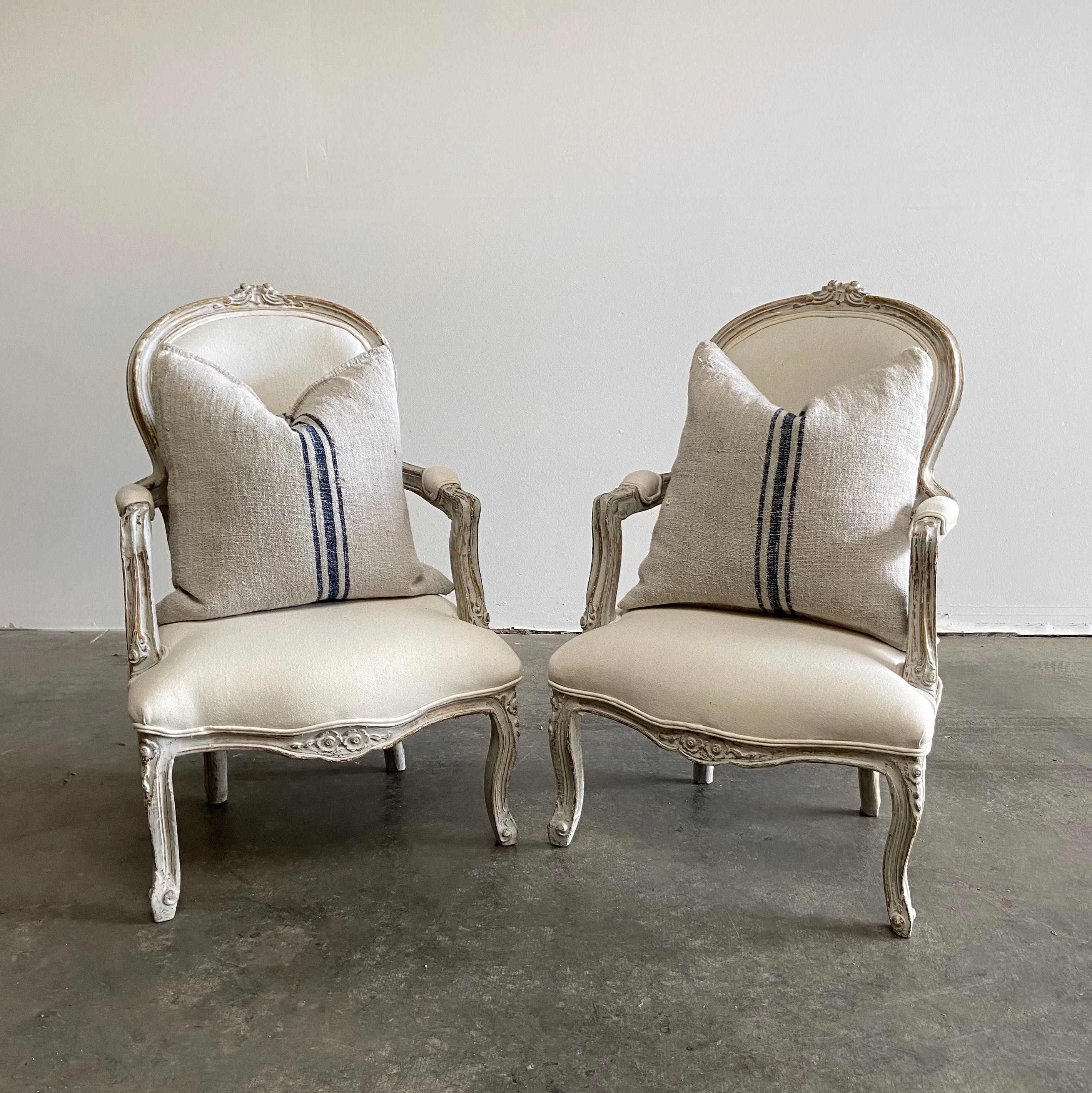 Pair of painted and upholstered Louis XV style open armchairs
Beautiful painted chairs in soft white with subtle antique distressing. An oyster grey hue, with gilt flecks peeking through the paint. Soft rounded scrolls, and hand carved flowers, and