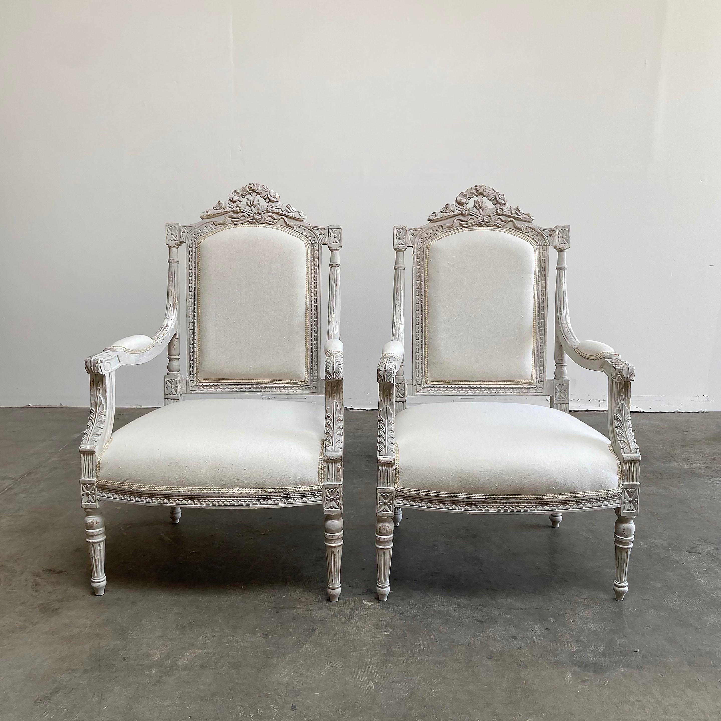 Vintage pair of painted and upholstered Louis XVI style chairs carved roses, flowers and scrolls across the top back, with a sleek fluted Louis XVI style leg. Painted in a layer of Gustavian gray, with antique white, and original gilt peeking