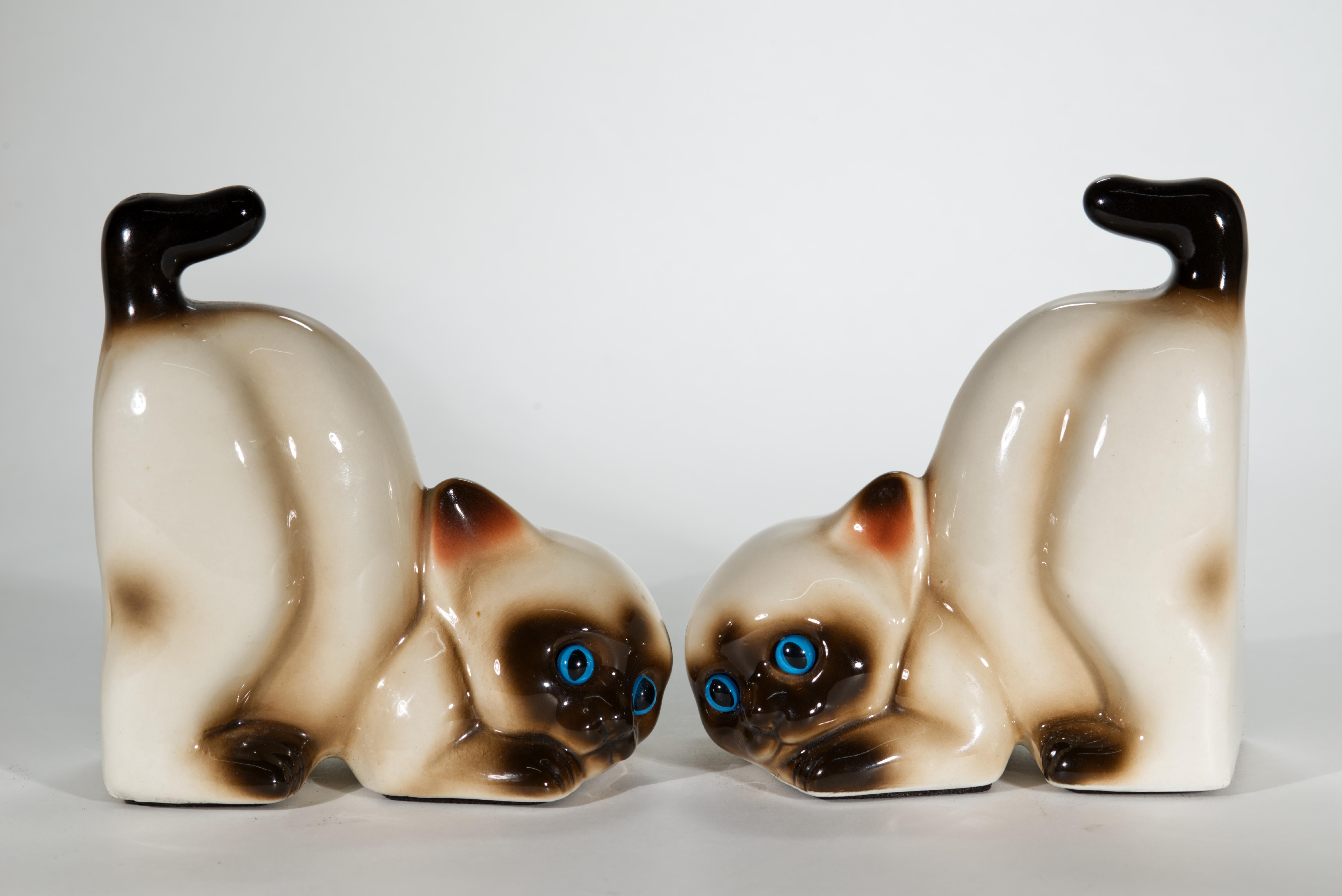 Questa adorabile coppia di fermalibri vintage presenta due giocosi gatti o gattini siamesi beige e marroni con luminosi occhi azzurri. I gatti sono scolpiti con corpi affusolati, code arricciate e sguardi maliziosi e leggermente sorpresi.

I gatti
