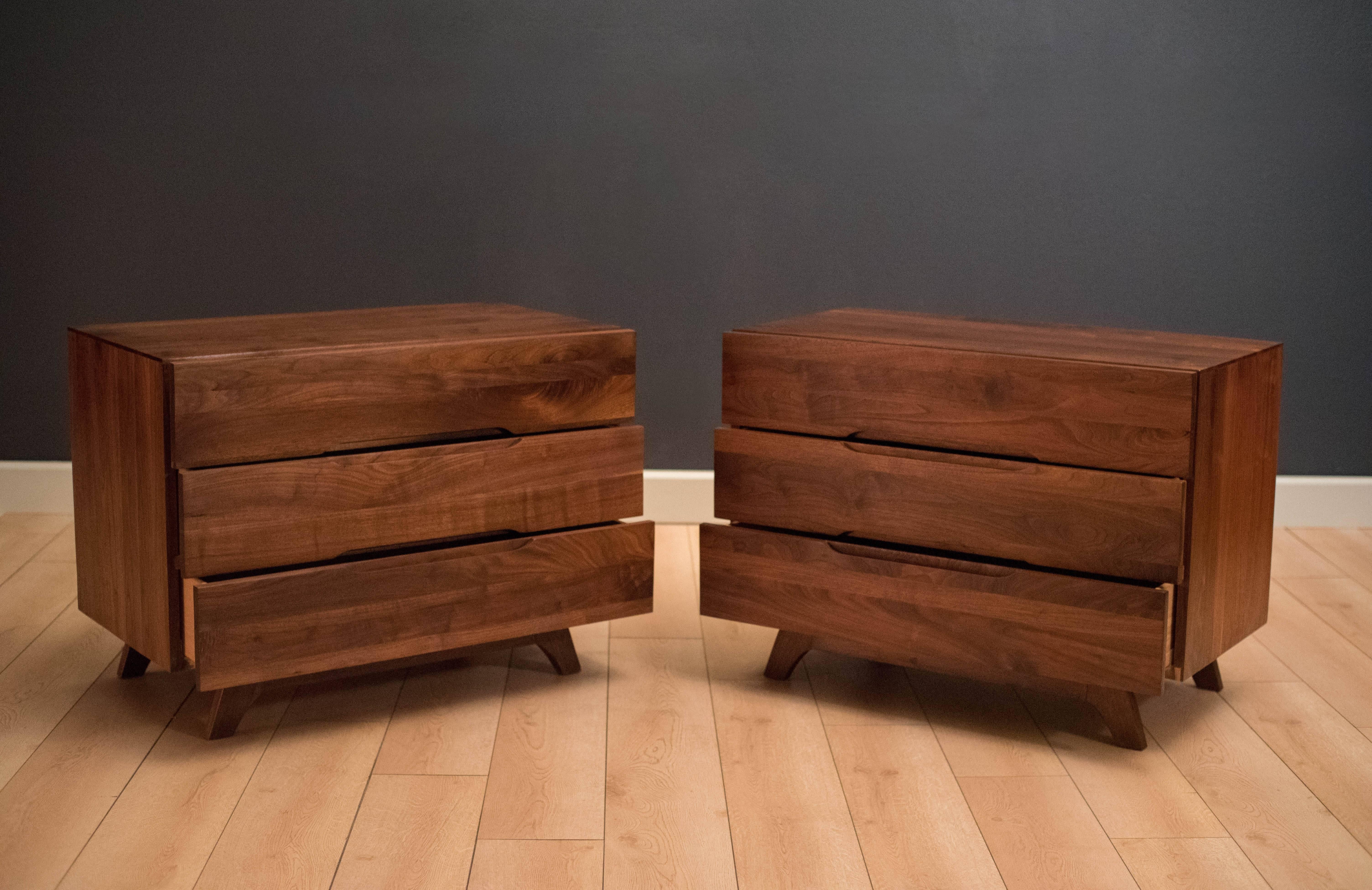 Mid-Century Modern pair of dresser chests or commodes made of solid planked walnut. Each chest includes three drawers with sculpted inset handles and a splayed leg base. 

 