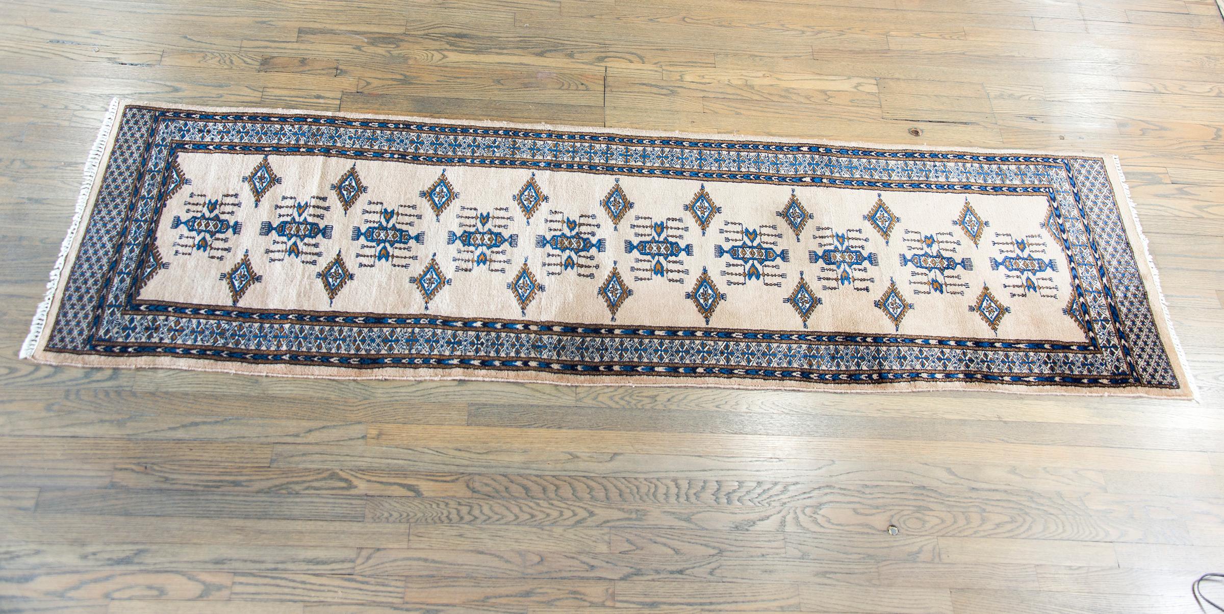 Un runner Bokhara pakistano della fine del XX secolo con molti grandi medaglioni a rombi e motivi floreali stilizzati ripetuti su un fondo color crema, circondato da un bordo complesso con diverse strisce stilizzate a motivi floreali e geometrici,