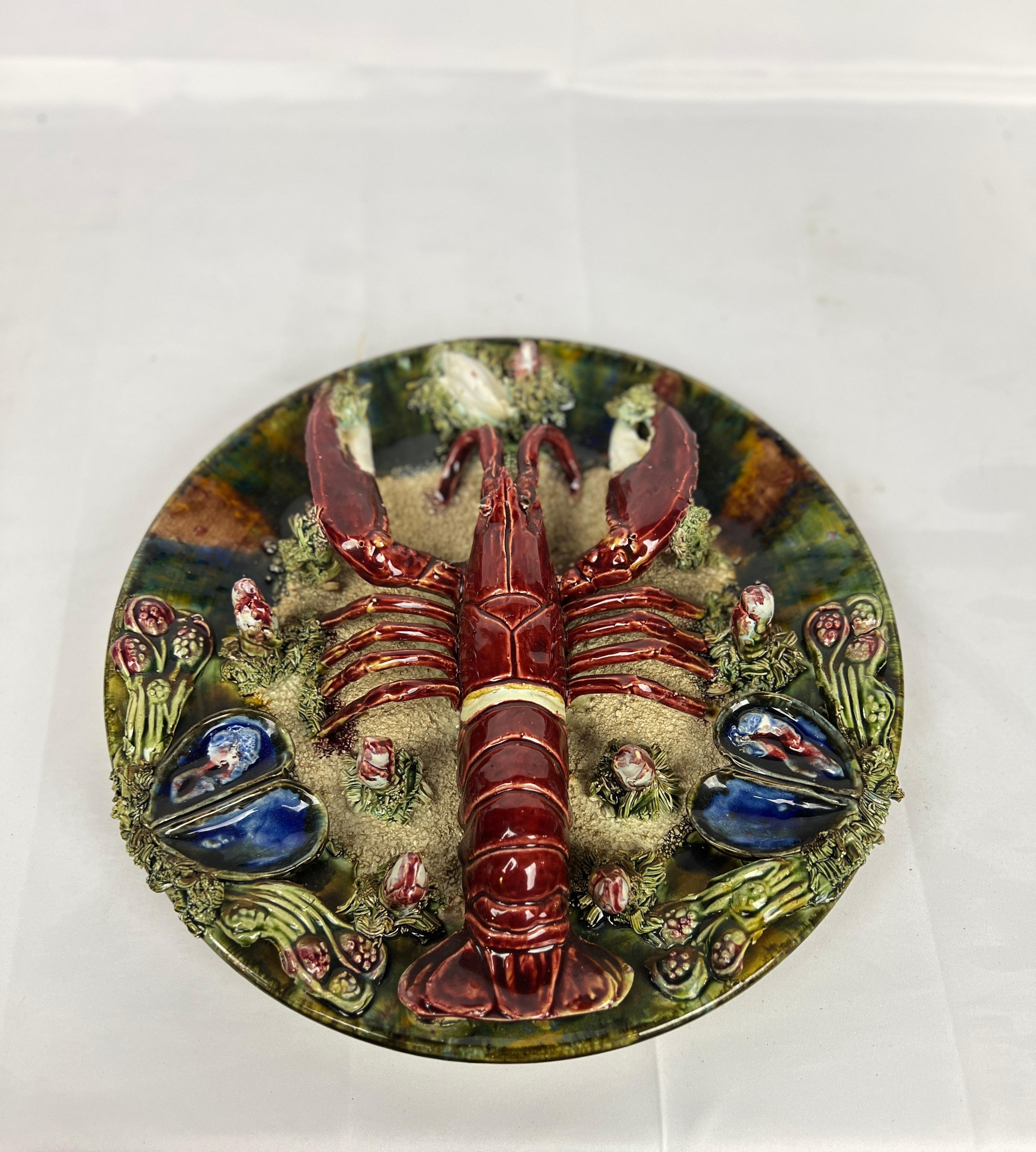 A vintage Palissy ware plate decorated with a central lobster figure which is surrounded by oyster and clam shells protruding from the edges. 

Measures: 12.375