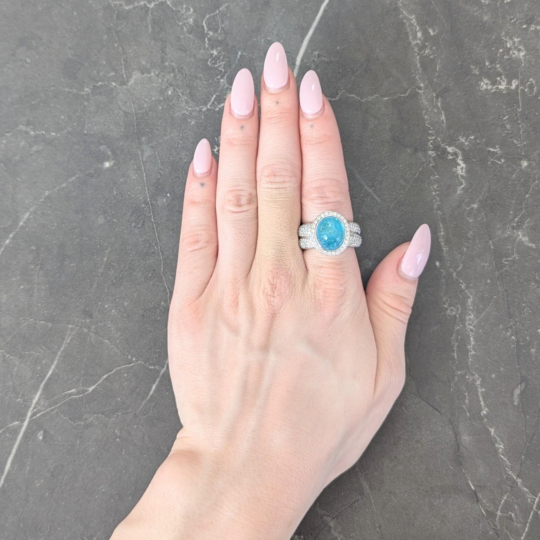 Centering an oval shaped tourmaline cabochon measuring 12.36 x 10.33 mm
Natural Mozambique in origin and displaying Paraiba properties
Translucent bright green-blue in color and bezel set
With a halo surround and puffed twist motif shoulders
Pavé