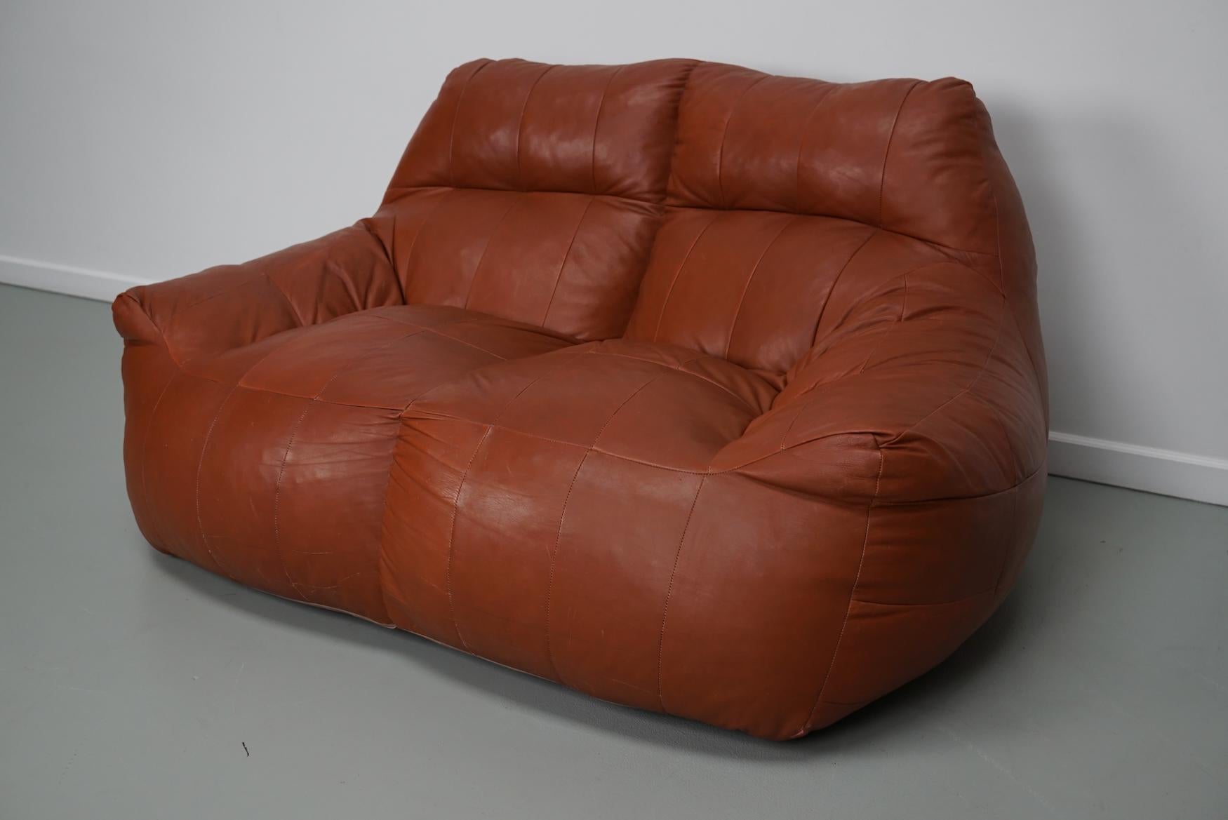 Ce confortable canapé vintage a été fabriqué vers les années 1970 en Allemagne. Il était recouvert de cuir patchwork marron/cognac et rempli de billes de polystyrène. Le canapé / pouf reste dans un bon état vintage avec seulement des signes minimes