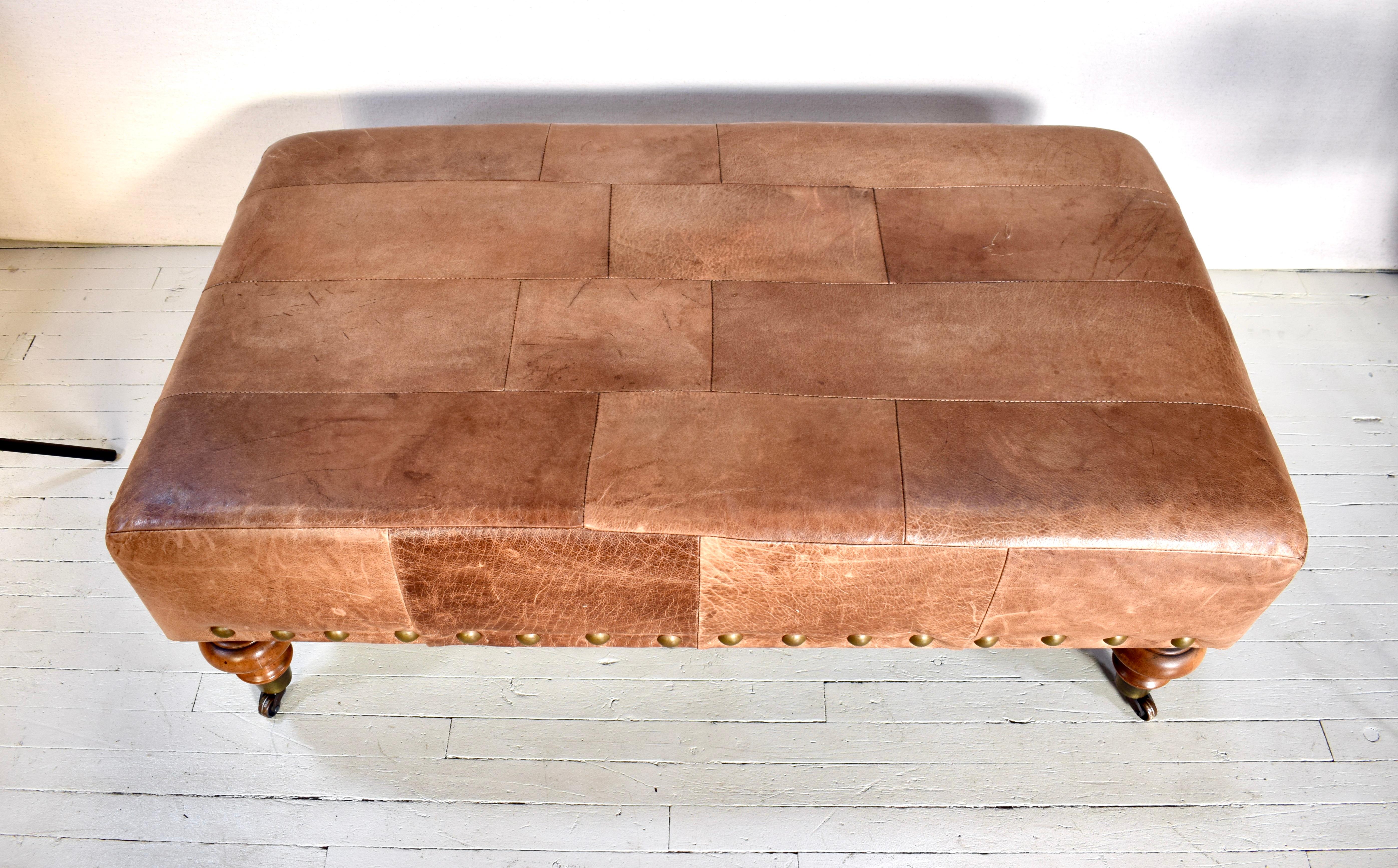 Vintage leather patchwork ottoman or bench embellished with large brass nail head trim on turned legs with brass casters. New Western in style with rich Carmel, Sienna & Amber tones; hand oiled boasting warm patina.