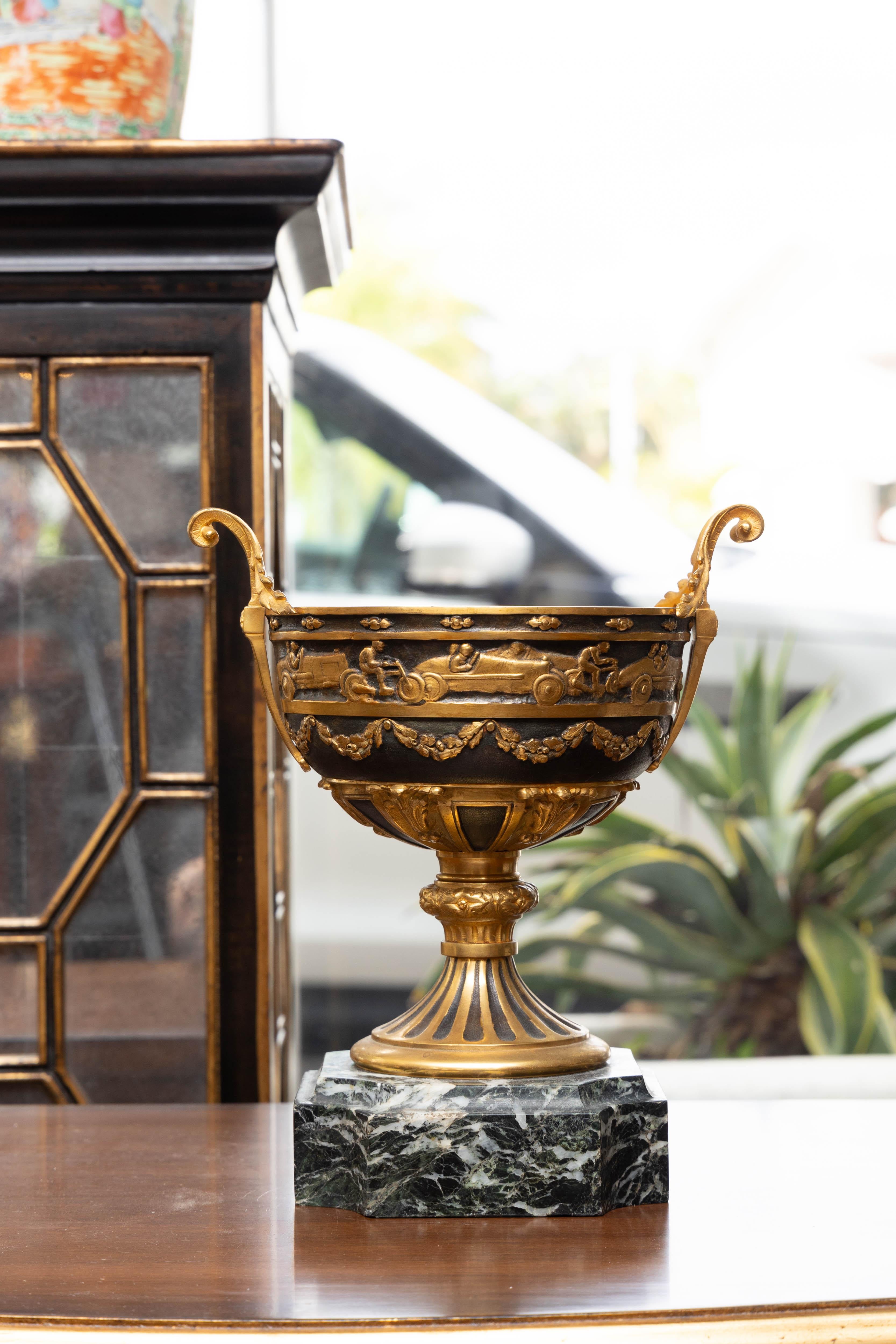 This is a stately looking patinated and gilt bronze chalice-style urn situated on a Verdi marble base.  This is possibly a trophy cup, featuring depictions of a variety of early race cars and motor cycles around the upper circumference and