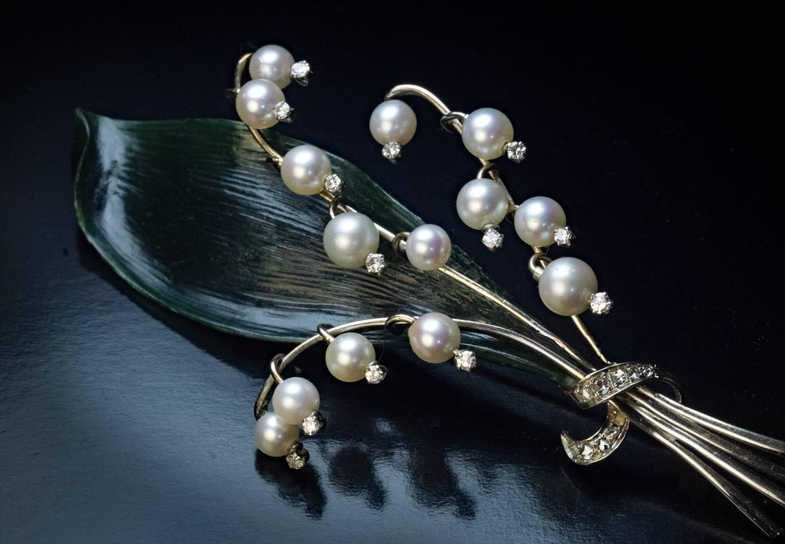 Made by prominent Austrian jeweller Jul. Hugler, Vienna, circa 1950.

The 18K white gold brooch is designed as a ribbon tied lily of the valley with forteen cultured pearl (5-6 mm) flowerheads accented by single cut diamonds laid on a