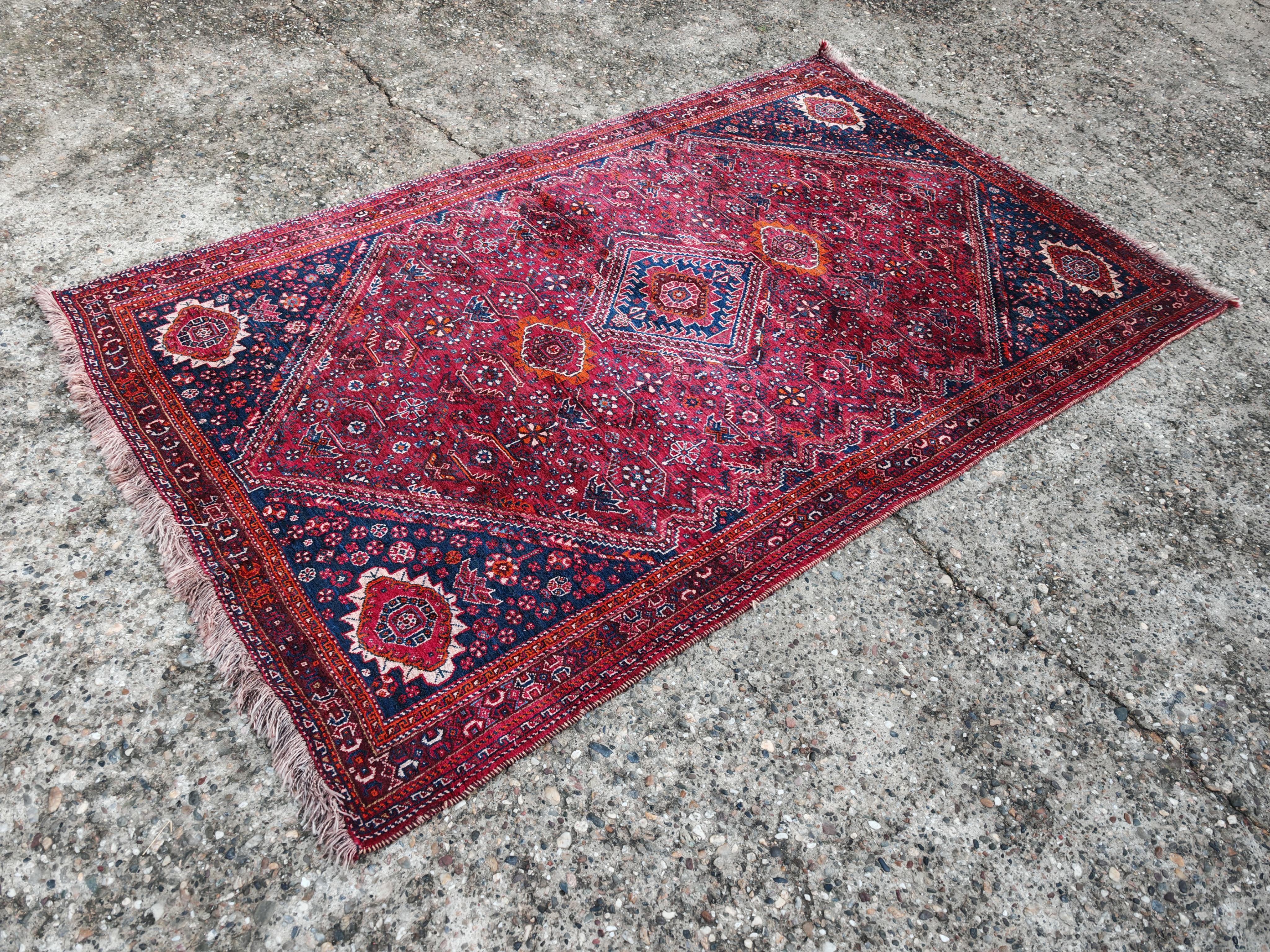 In questa inserzione troverai un grande tappeto tribale persiano vintage annodato a mano. Il colore rosso intenso del campo, i bordi blu navy e i disegni geometrici con motivi ottagonali o a rombi che si ripetono sono tipici di questi stili