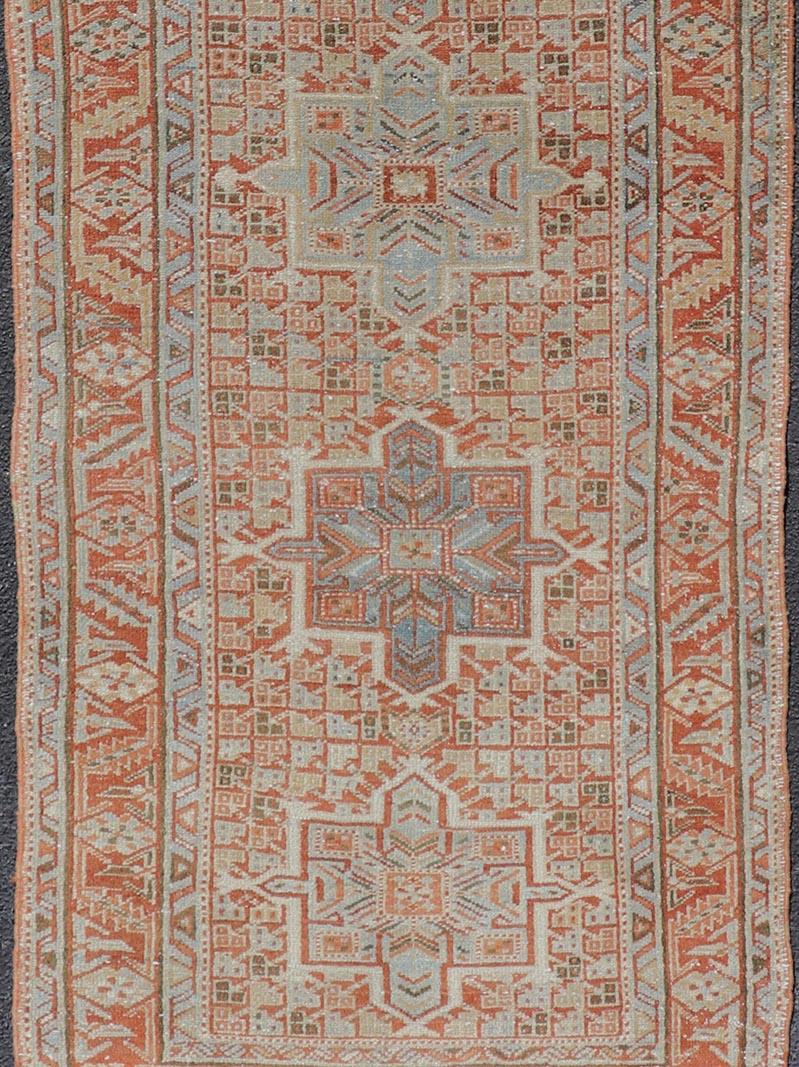 This antique Persian Heriz rug features a distressed all-over sub-geometric design rendered in soft red as well as light blue tones, and is set upon an ivory background. A complementary, multi-tiered border encompasses the entirety of the piece;