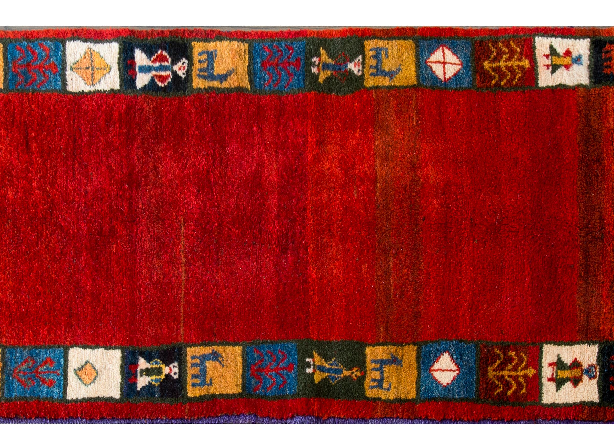A gorgeous mid-20th century Persian Gabbeh rug with a bold abrash crimson field surrounded by myriad squares with stylized goats, people and trees-of-life woven in gold, indigo, green, and crimson.