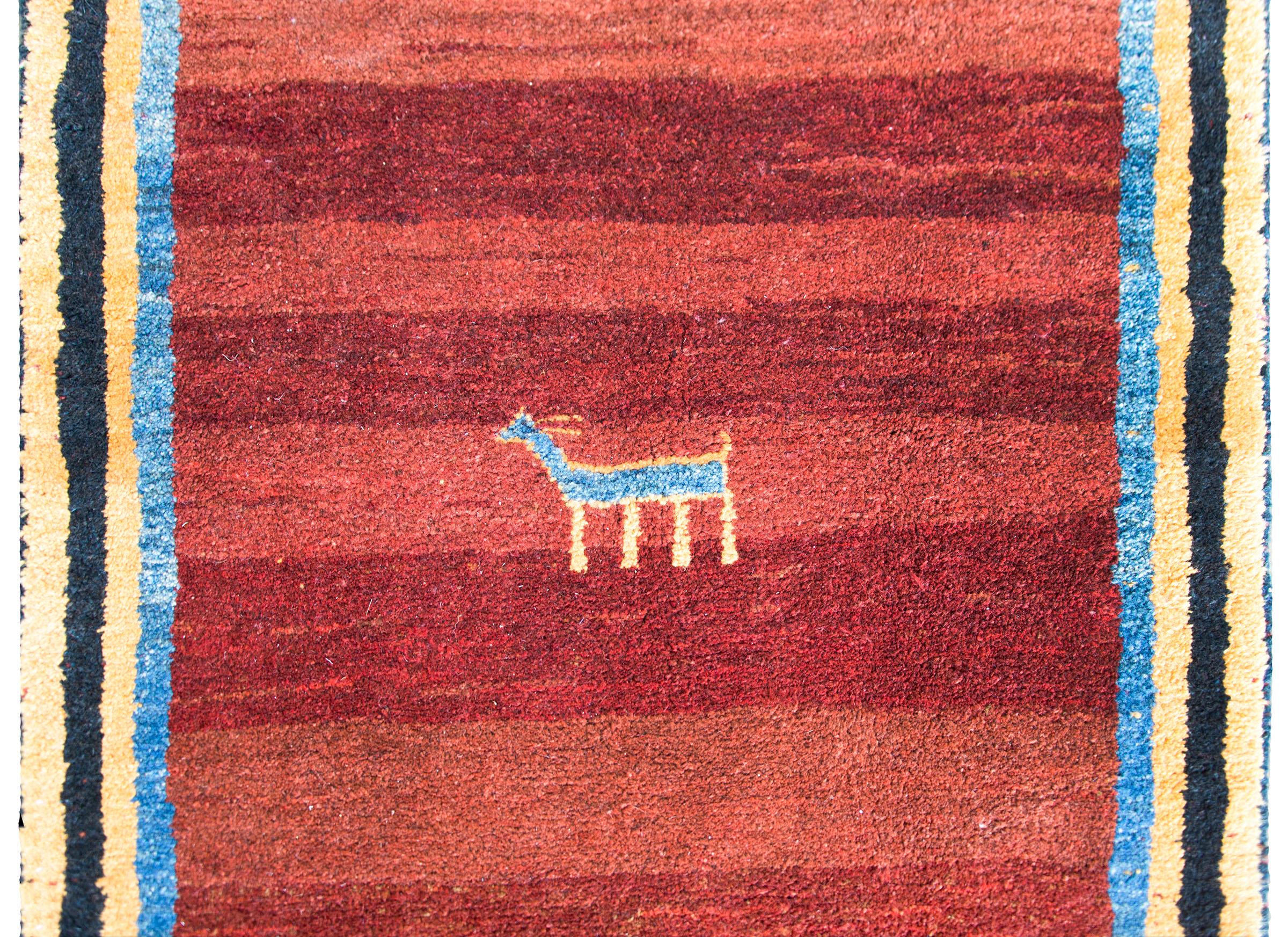 A bold vintage Persian Gabbeh rug with a abrash cranberry field with a central stylized indigo goat, more goats and people at each end, and surrounded by a black, gold, and indigo striped border.