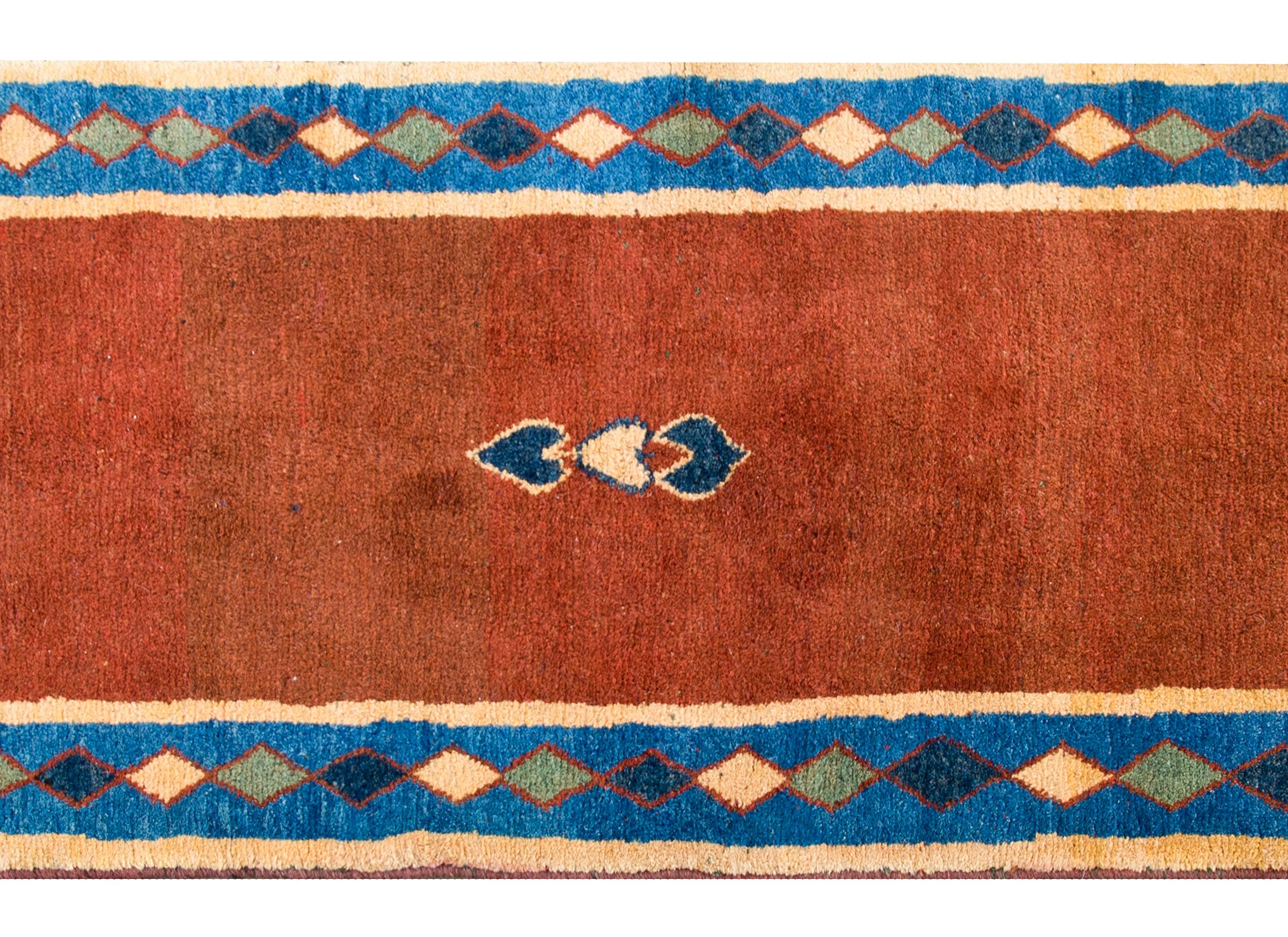 A fantastic mid-20th century Persian Gabbeh rug with a burnt orange field with three small stylized leaves in the center, and surrounded by a harlequin diamond border woven in indigo, gold, green, and a thin red line stripe.
