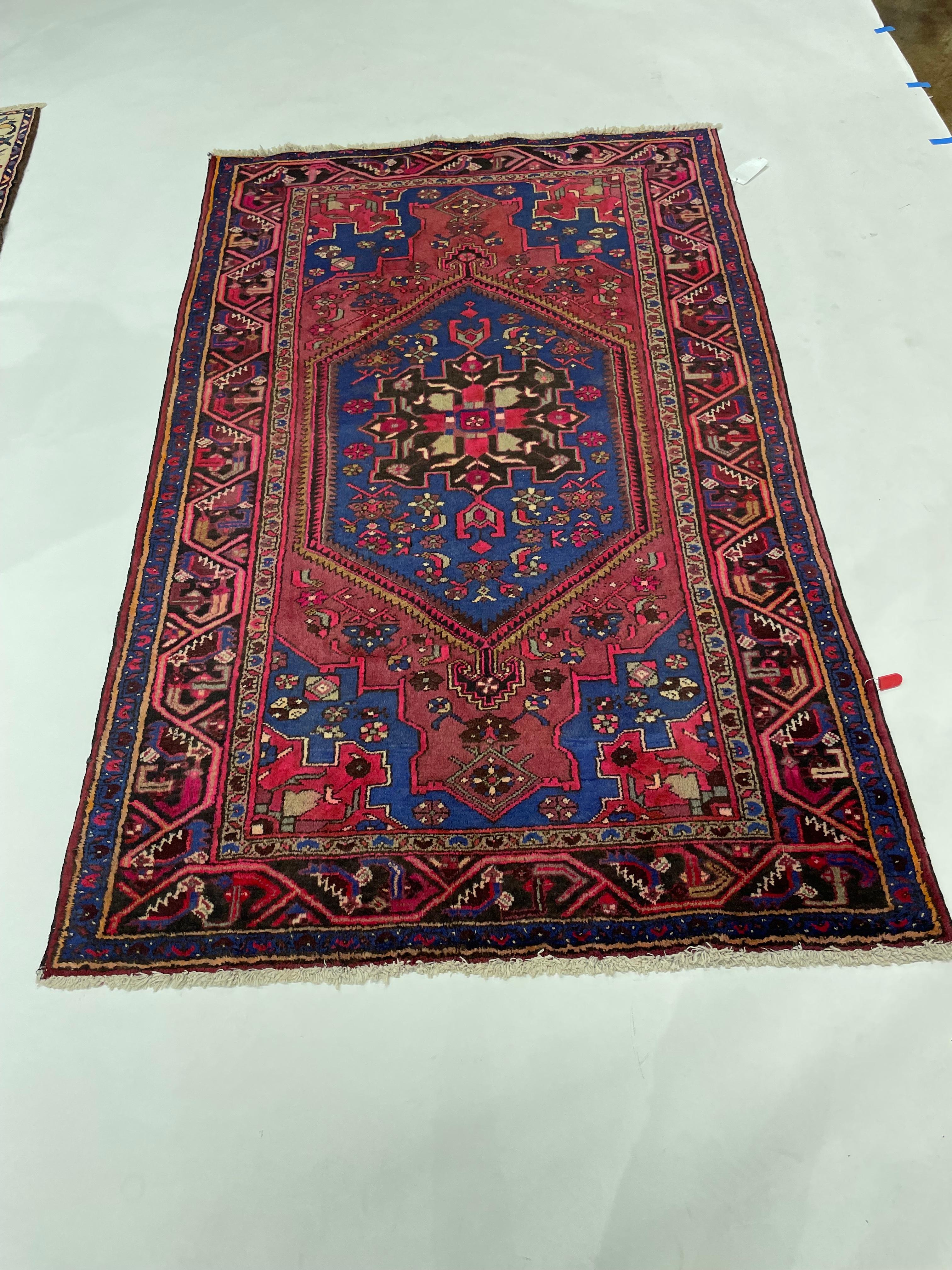 Dieser auffällige handgeknüpfte Perserteppich zeigt ein kühnes geometrisches Medaillon in einem leuchtend kobaltblauen Feld, das von einem satten Preiselbeerrot umrahmt wird. Das Design ist mit stilisierten Blumenmotiven, Tieren und charmanten