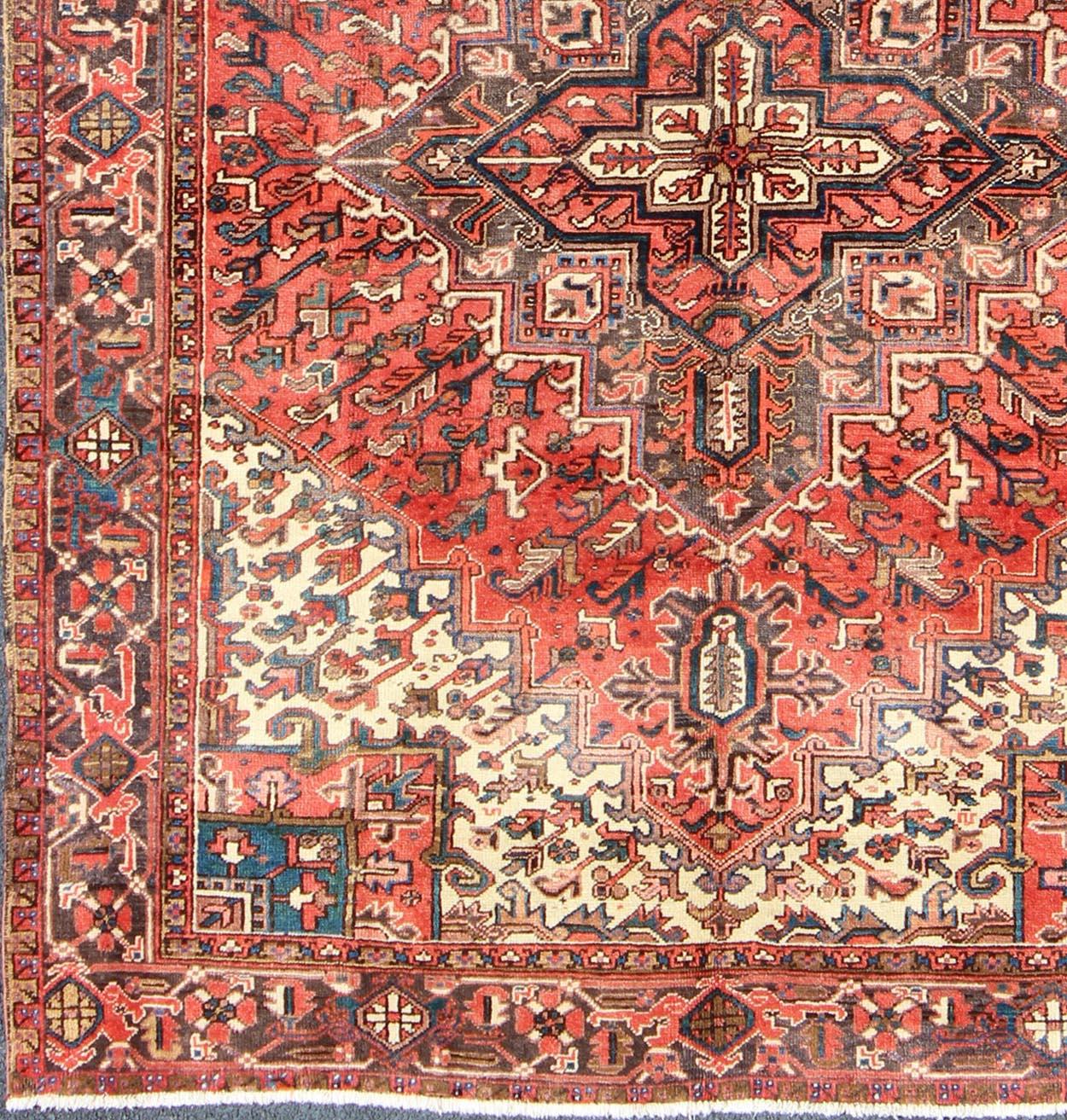 This magnificent antique Persian Heriz from the Mid-20th century bears an exquisite design rendered in gorgeous, warm hues of rust and denim blue. A highly stylized geometric center medallion anchors the rug and is surrounded by numerous geometric