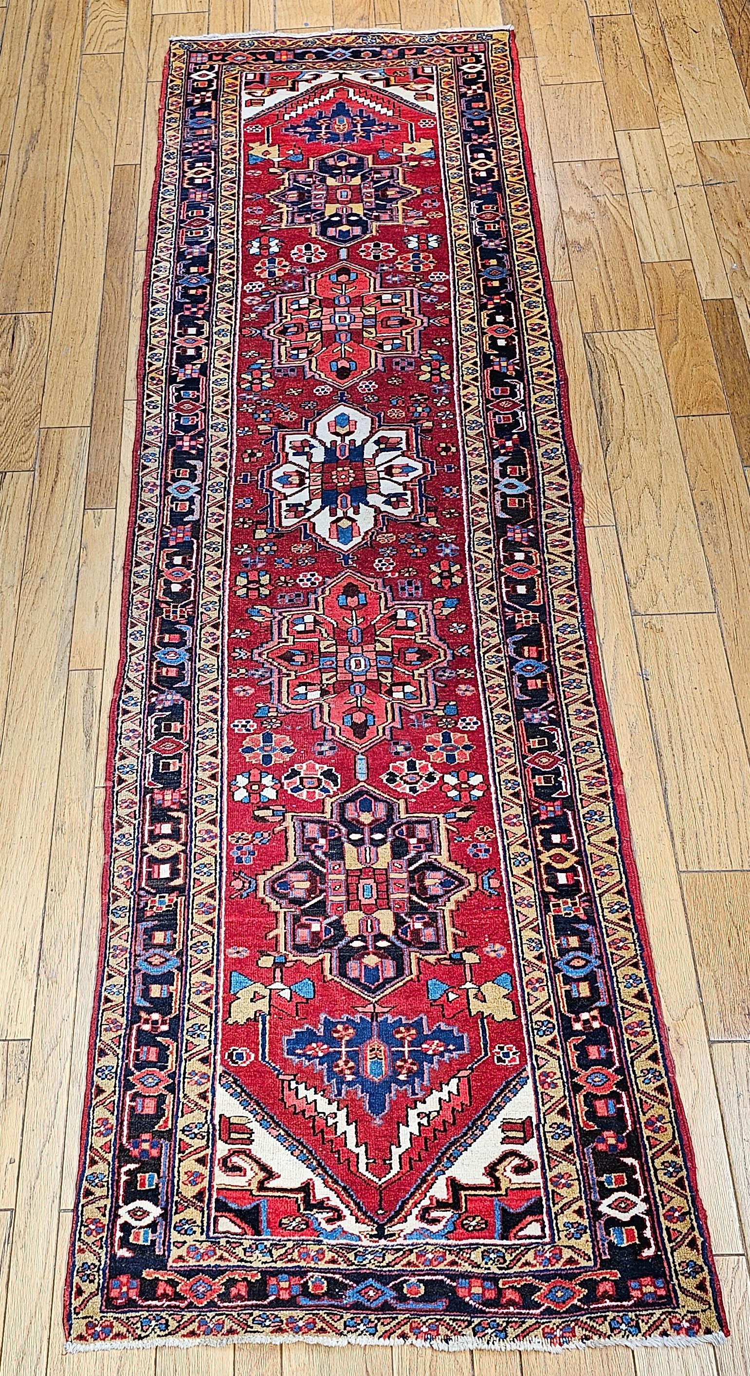 Dieser lebhafte persische Heriz-Läufer im Vintage-Stil zeichnet sich durch eine auffällige geometrische Komposition mit fünf axialen Medaillons in tiefem Marineblau, Elfenbein und sattem Rot aus, die alle vor einem leuchtend roten Feld stehen. Das