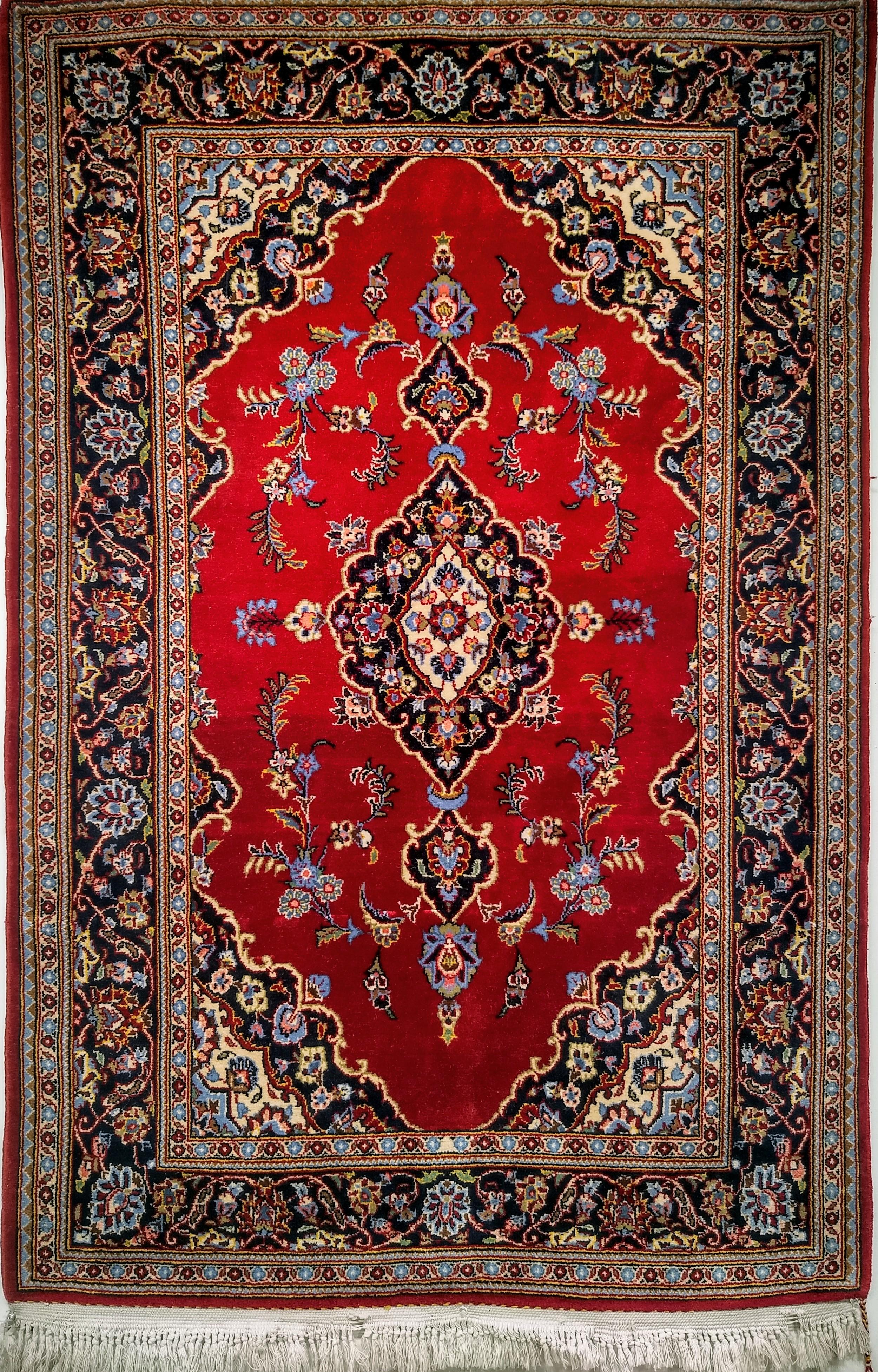 Alfombra persa vintage de Kashan con un clásico motivo floral en medallón de mediados del siglo XX. La alfombra presenta un hermoso campo de color rojo intenso con un medallón central y un borde azul oscuro. El sencillo diseño de campo despejado de