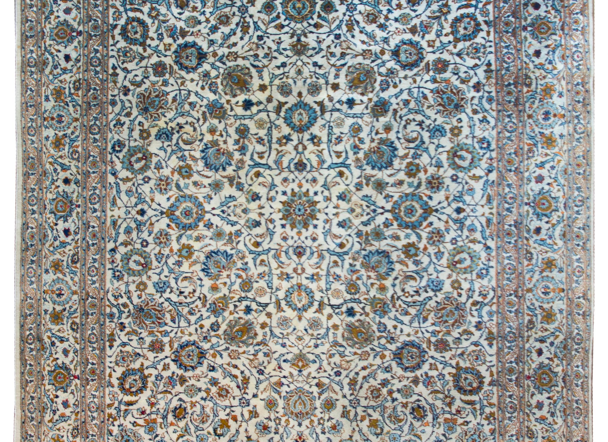 A beautiful mid-20th century Persian Kashan rug with an all-over intensely woven floral and scrolling vine pattern with myriad flowers woven in light and dark indigo, crimson, and copper colored wool, set against a cream colored background, and