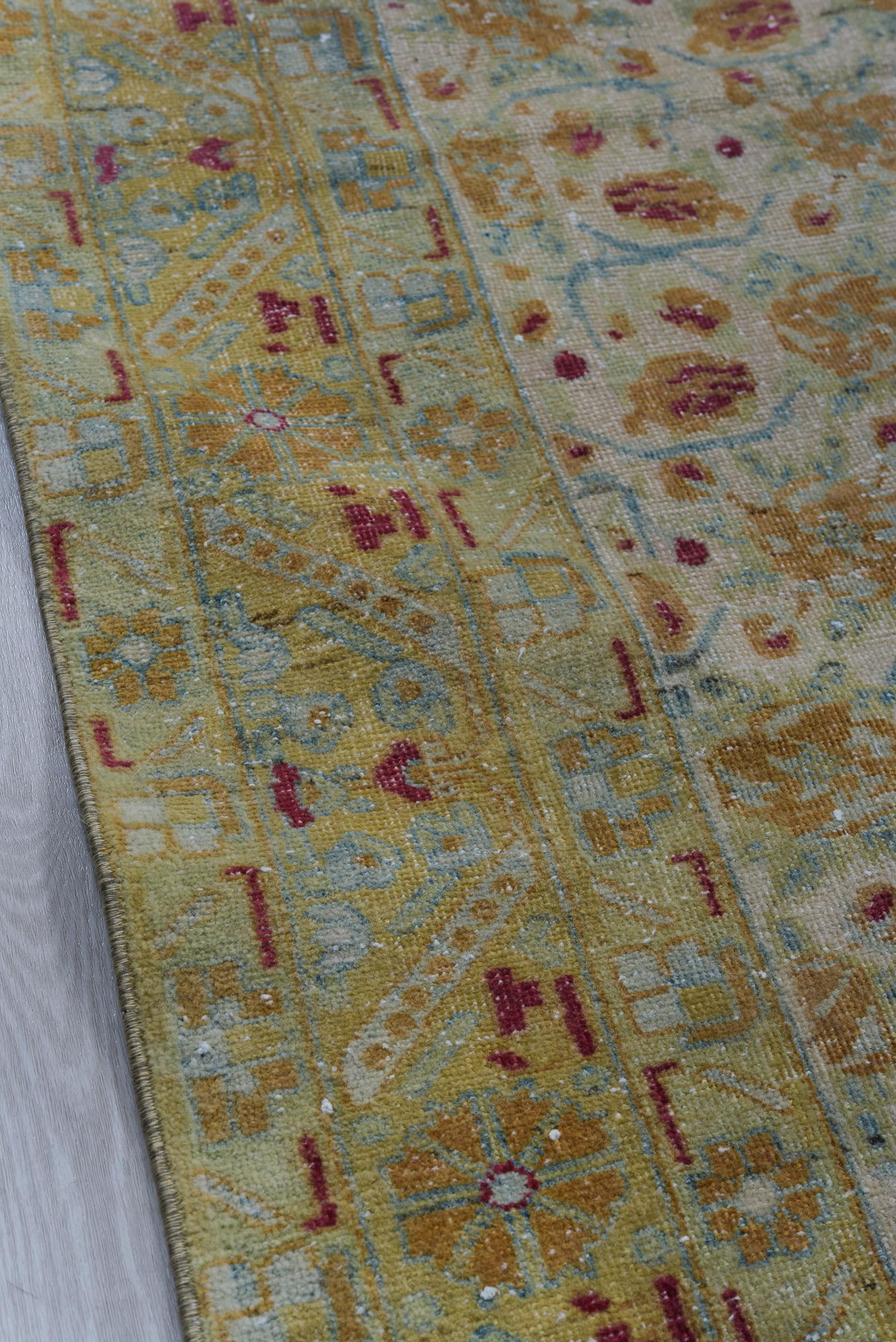 This vintage Persian rug features a delicately aged celadon field adorned with a dense rosette repeat in ochre, blush, and soft crimson. The floral pattern is framed by an intricate golden border filled with stylized blossoms and vinework in coral,