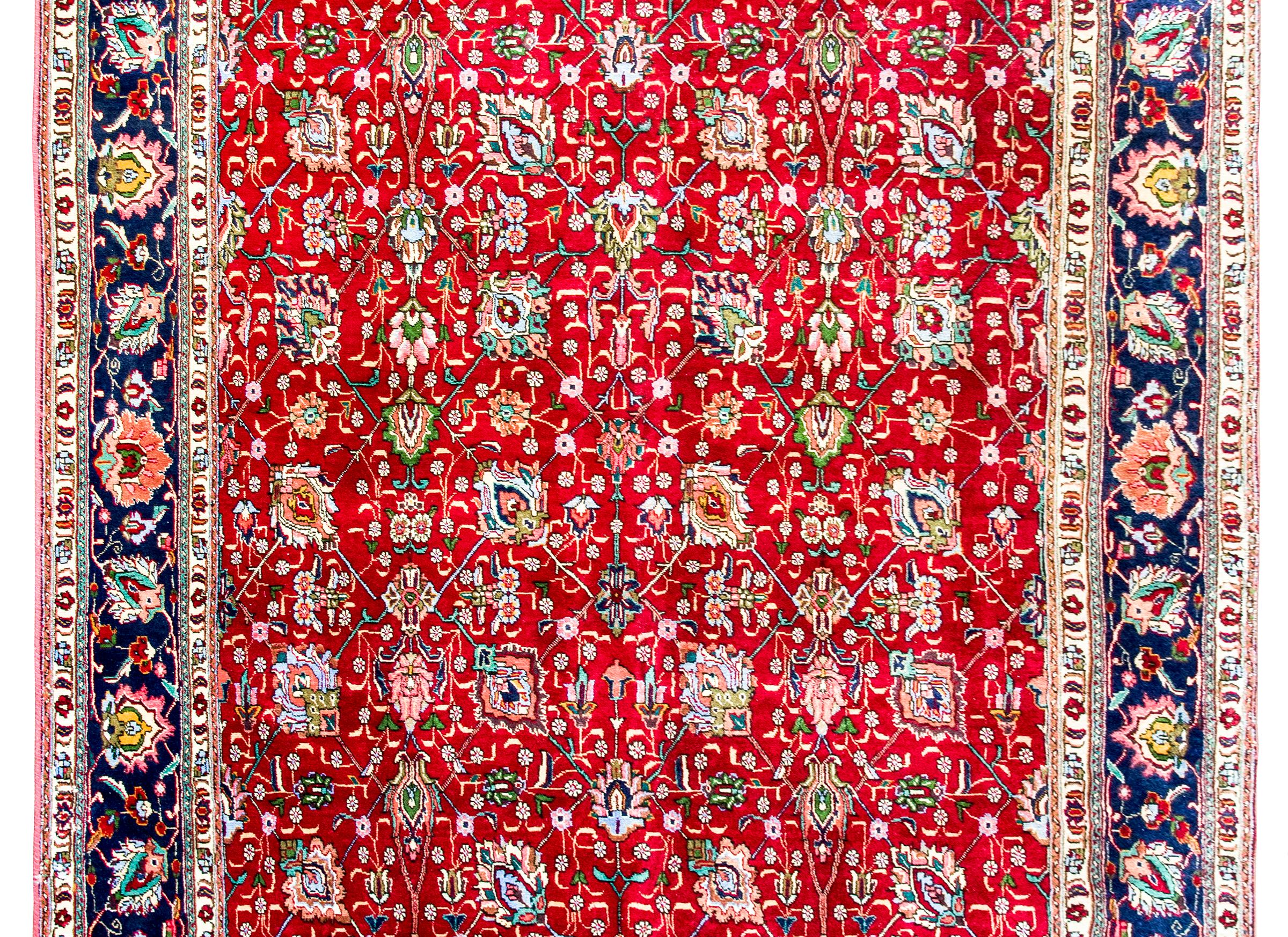 A wonderful mid-20th century Persian Tabriz rug with a large-scale repeated floral pattern woven in myriad colors including pink, green, orange, and light and dark indigo, and all set against a crimson background.  The border is beautiful with a