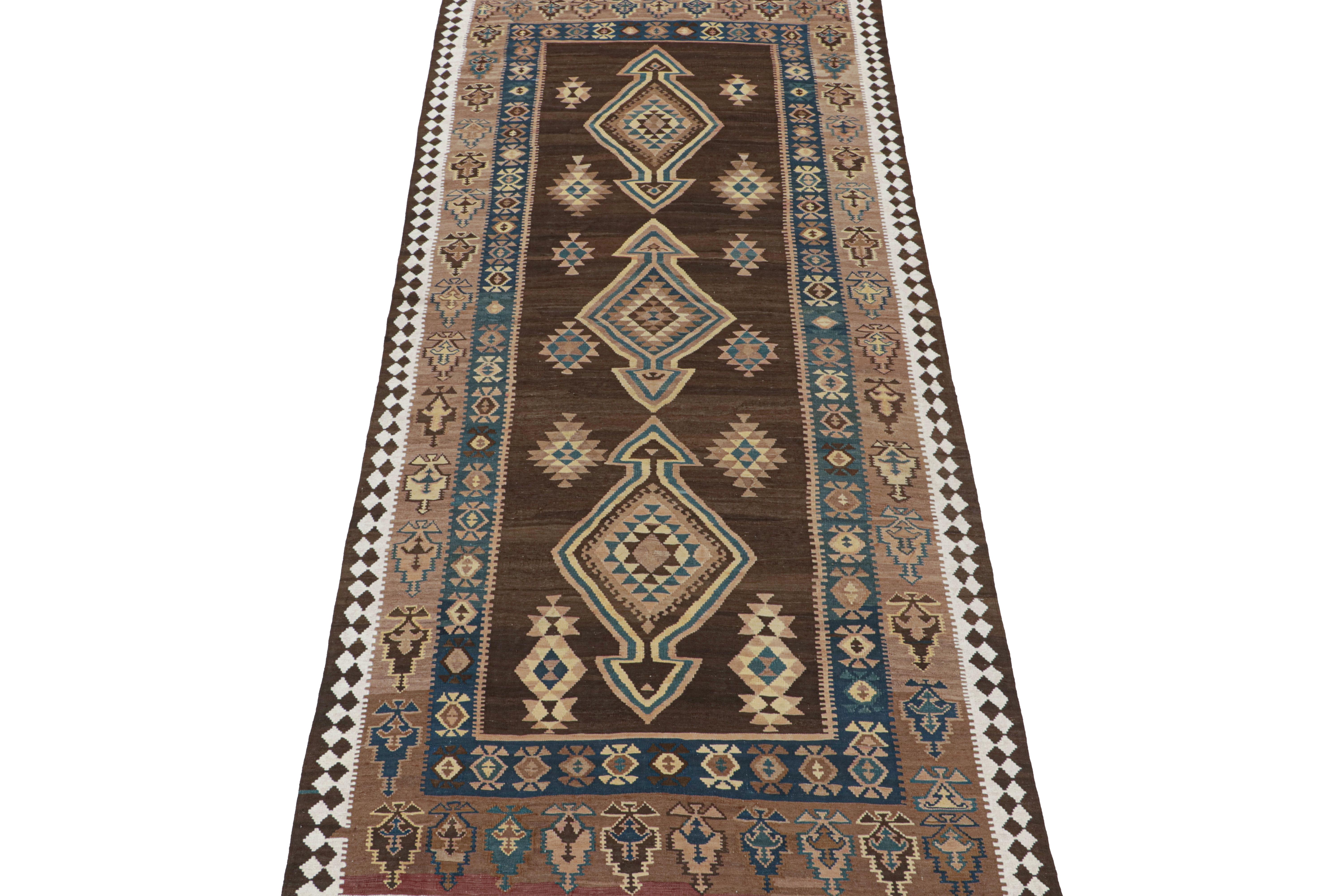 This vintage 5x13 Persian Kilim is believed to be a tribal rug of the 1950s. 

Handwoven in wool, its design enjoys medallions and geometric motifs in rich brown, beige, and blue tones. Connoisseurs will note the white fencepost-style border, and