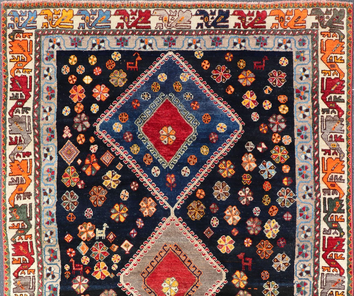 This vintage Persian Shiraz features a rich navy blue background under the centered red medallions, edged with green, gold, taupe, maroon, pinks and reds. Paired with earthy tones, such as greens, creams, oranges, and reds, in an intricate tribal