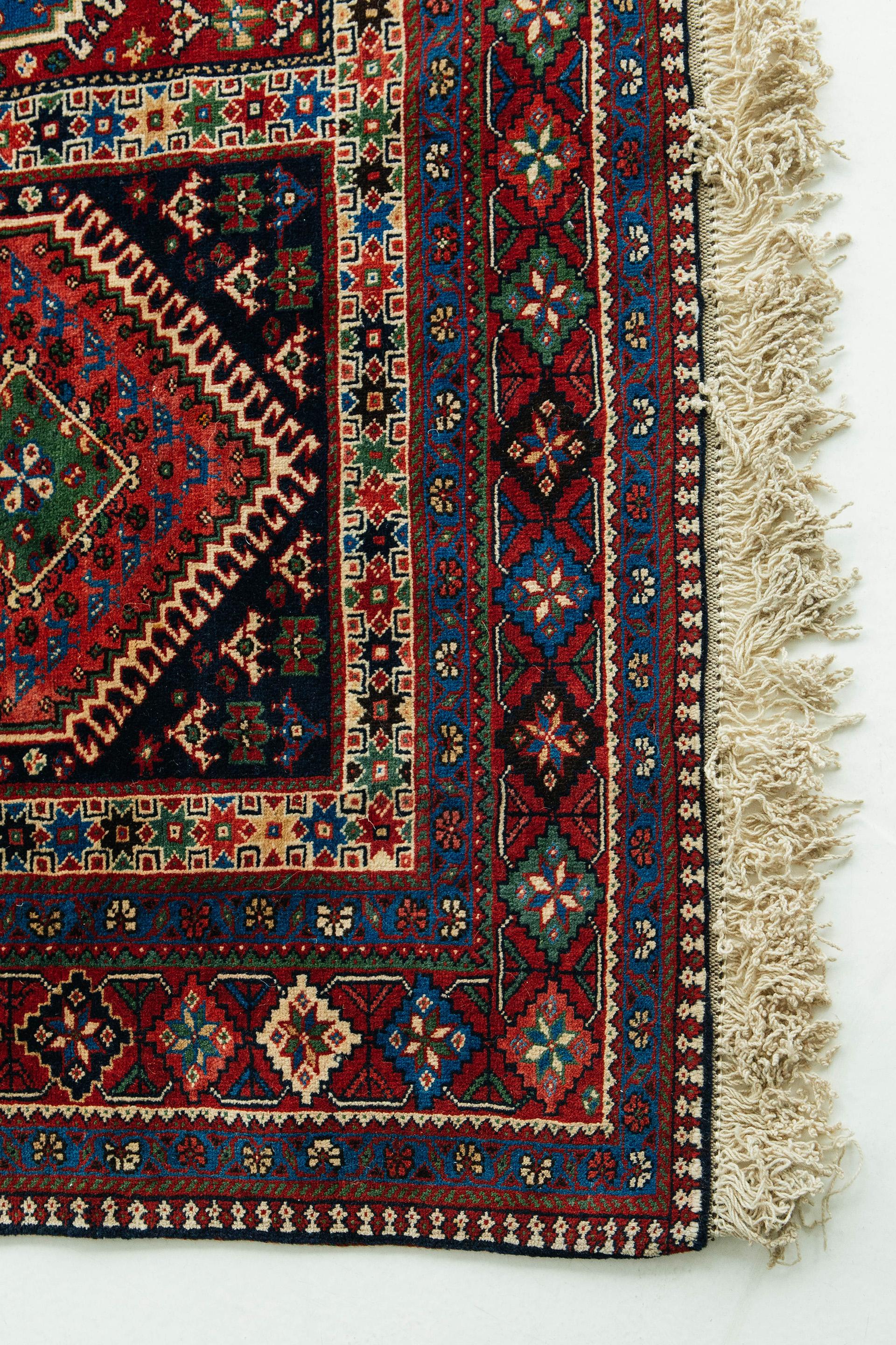 This antique Yalameh rug in deep red, blue, green, black, and ivory tones. The field pattern consists of rectangular compartments with latch-hook diamond medallions and varied fill motifs, including stars and talismans. The main border features