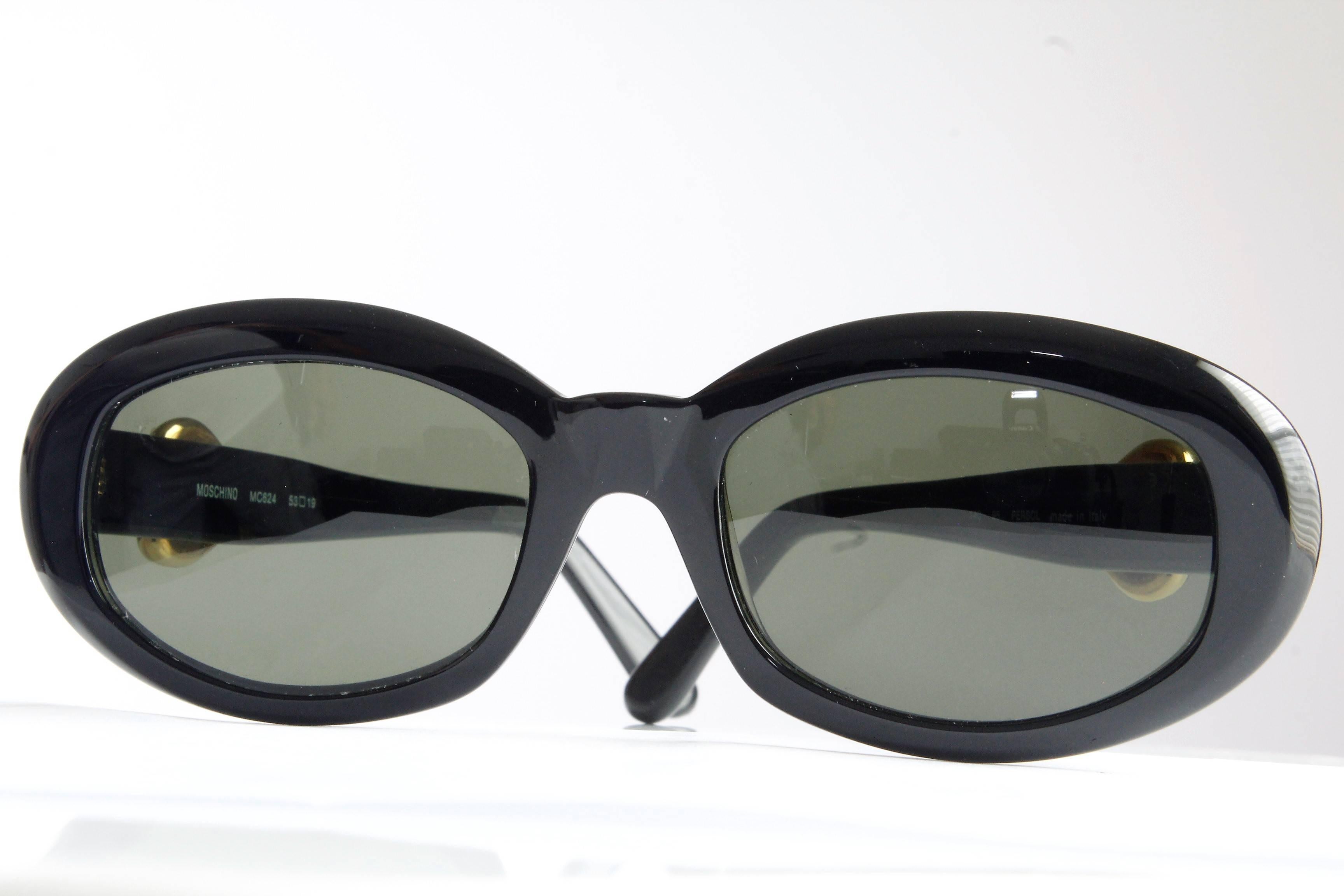 Paire de lunettes de soleil de forme ovale pour femmes, fabriquées par Persol Ratti Italy pour Moschino, vers les années 80.  

Fabriquées en résine noire, ces lunettes de soleil chics sont ornées de cœurs en métal doré de chaque côté.  

De qualité