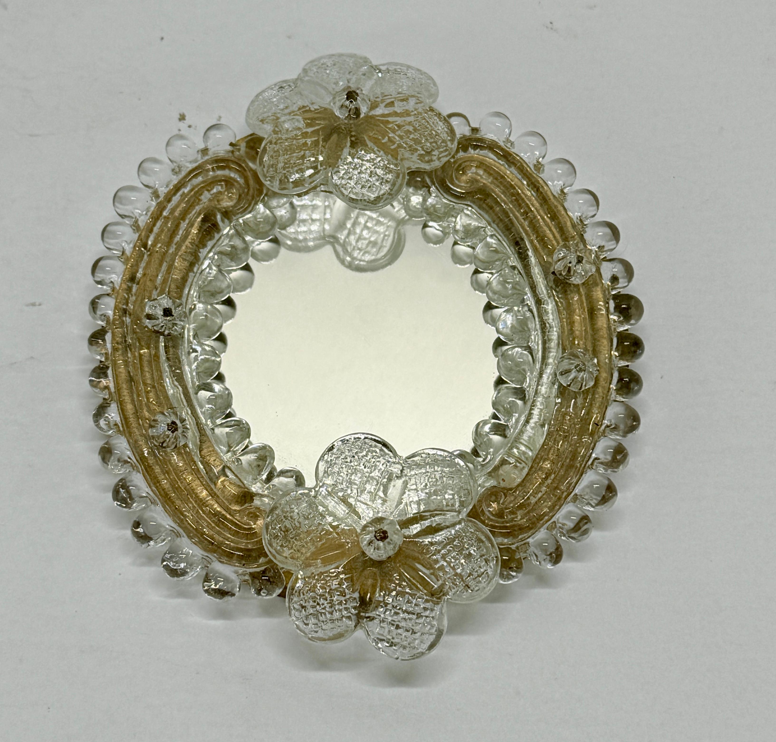 Un piccolo specchio in vetro artistico realizzato con due fiocchi di vetro veneziano rosa montati intorno a una cornice con bordi smerlati color oro. Verso è dotato di un gancio per l'utilizzo a parete. Adesivo di Murano sul verso.

Misure:
4,875