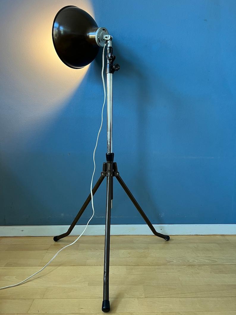 Vintage photography floor lamp. The lamps has a tripod-style base, similar to the stands used for old cameras and studio lighting equipment. The lamp has an adjustable height feature, allowing you to raise or lower the lamp to your desired level.