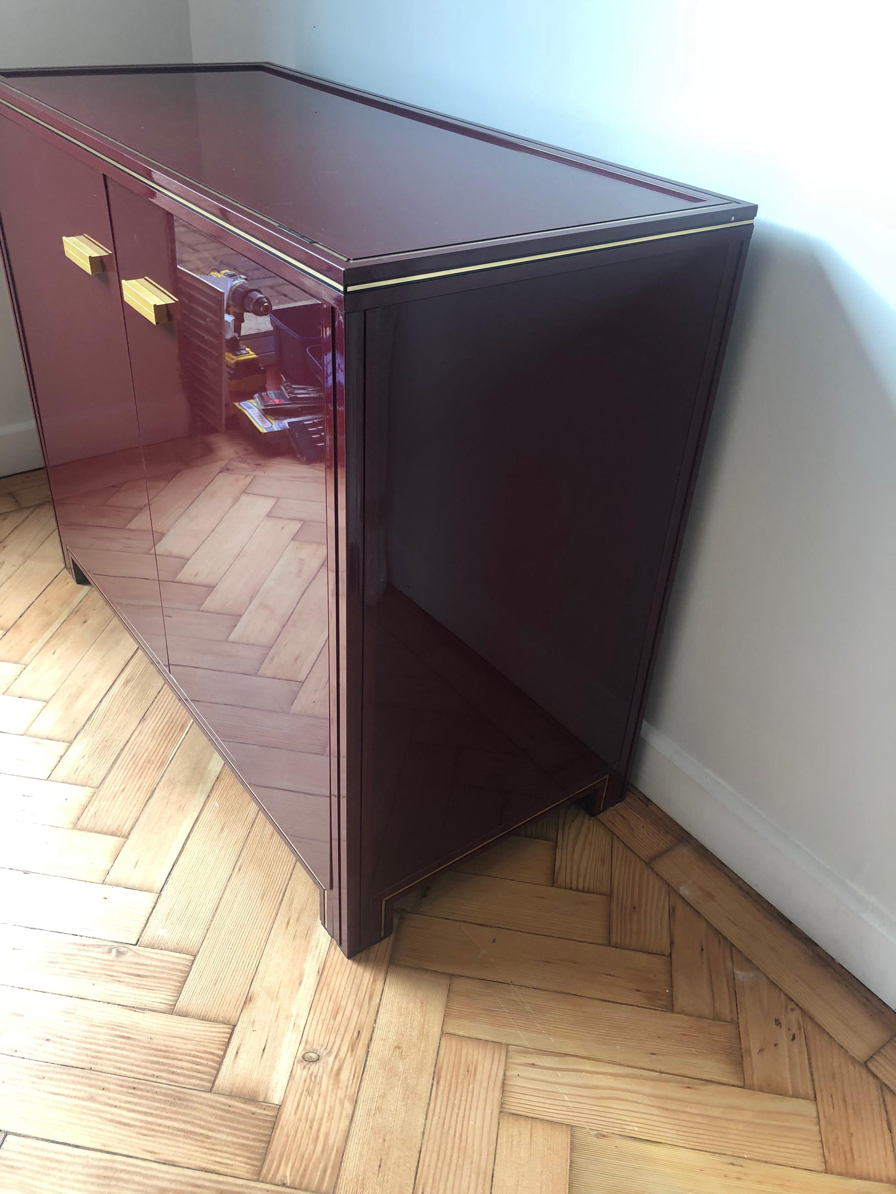 Pierre Vandel two-door cabinet in burgundy color with brass details, glass top, acrylic body and aluminum frame. The two doors open smoothly and it has a good storage size.
Overall good original condition with the occasional wear. 
 